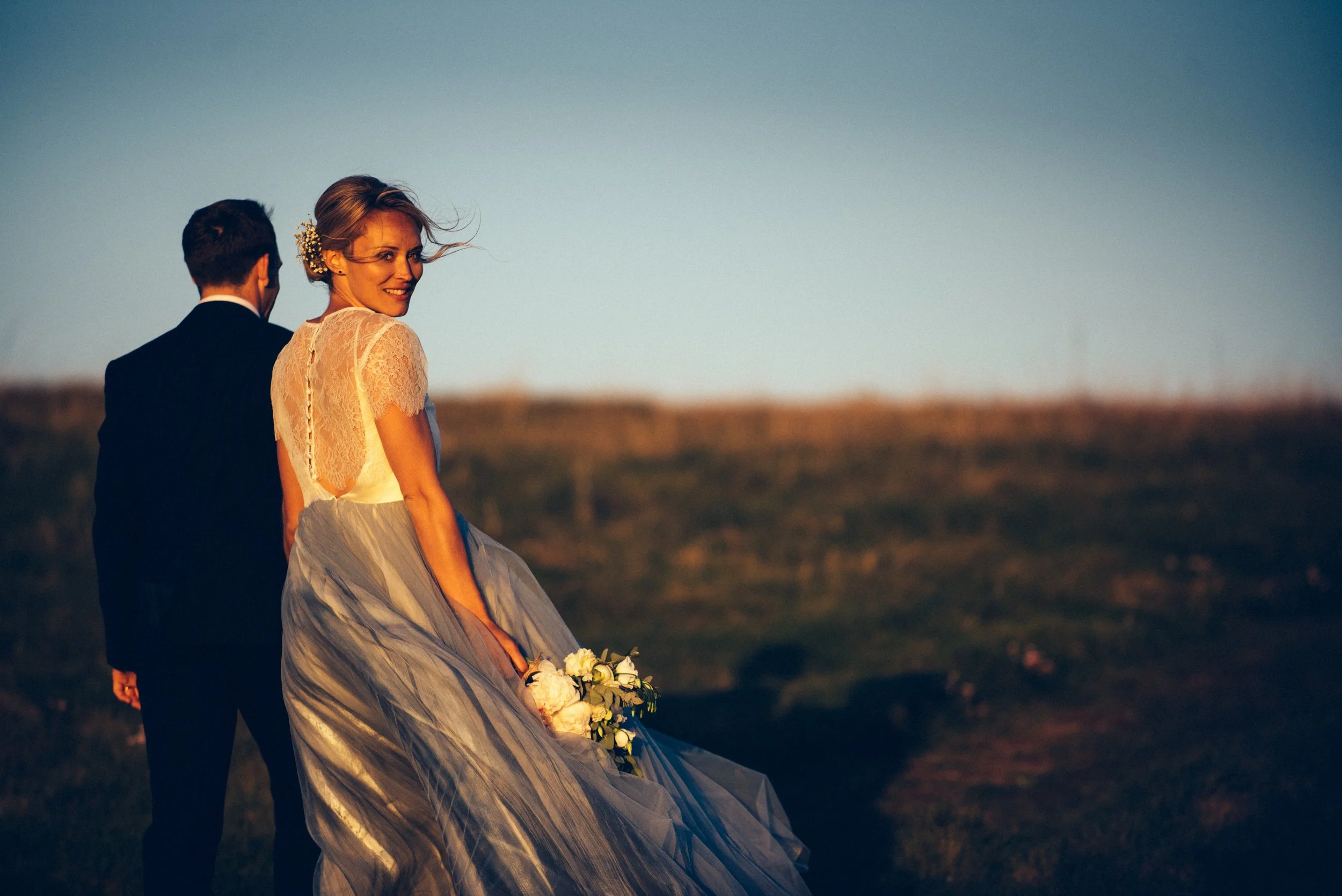 Bride in a lace wedding dress holding flowers, walking with a man in a suit during sunset, in a field background.