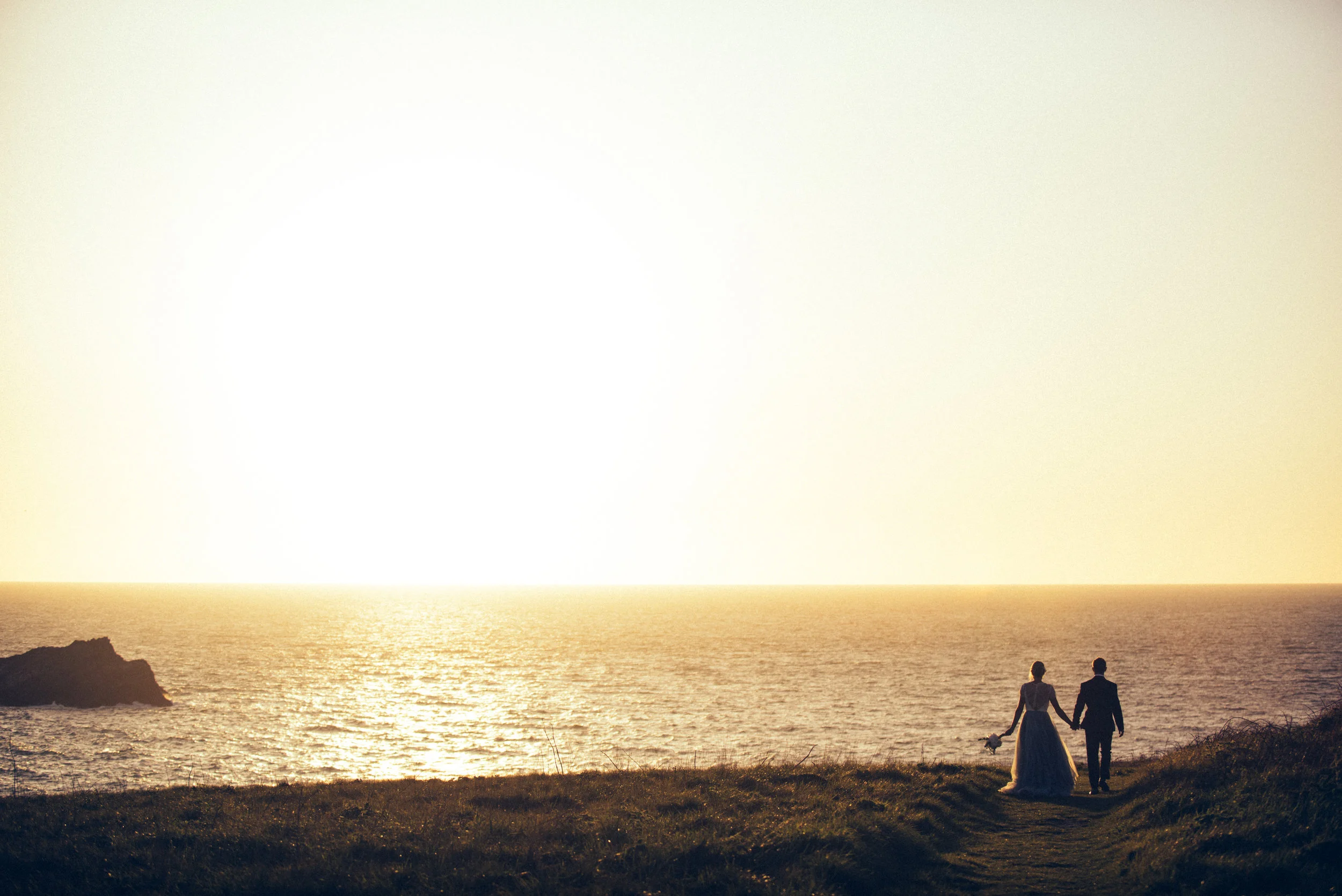Silhouette of wedding couple holding hands walking towards the sea at sunset. Photo taken at Crantock Bay on the north coast of Cornwall. 