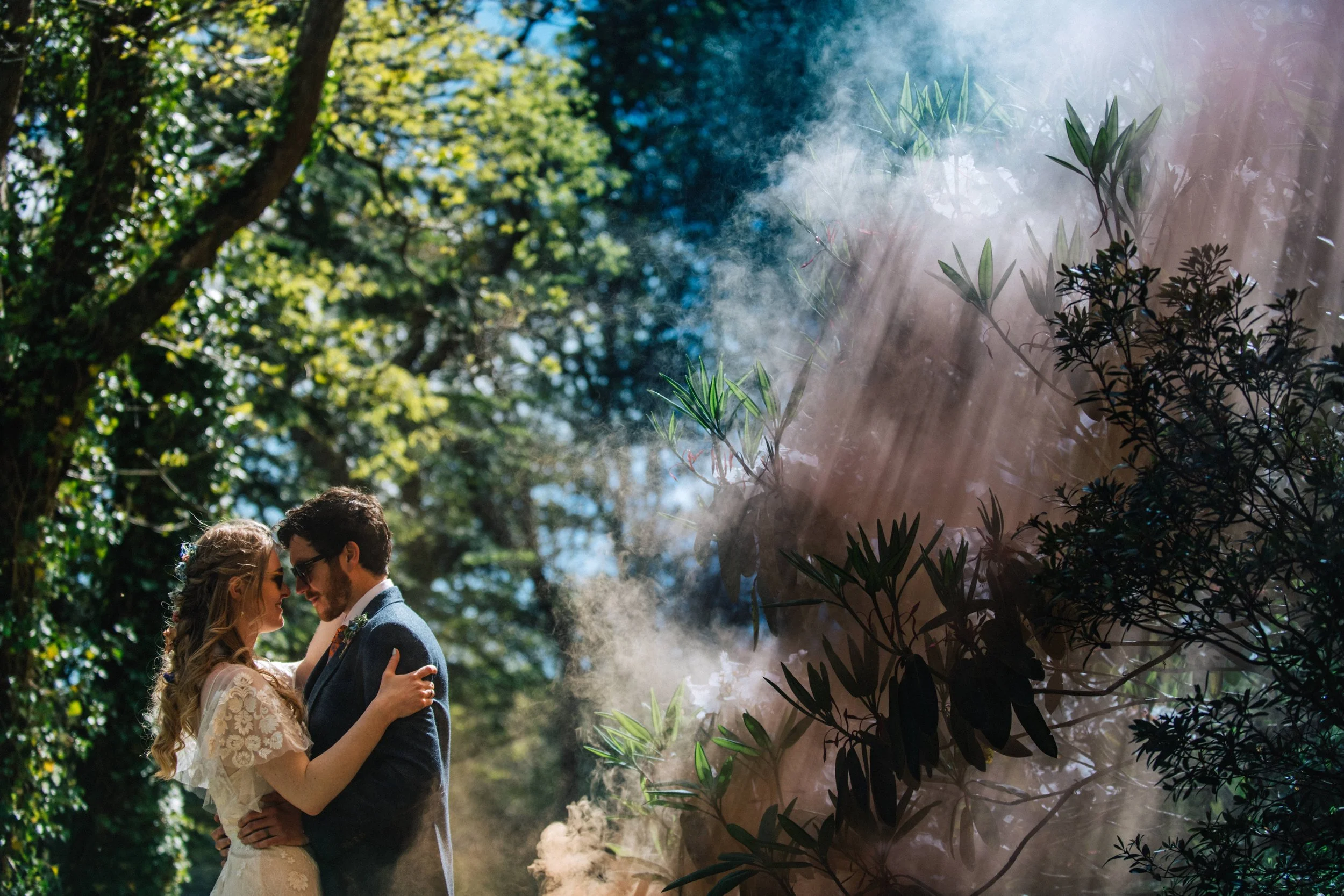 Couple embracing outdoors with sun rays shining through trees and smoke, romantic atmosphere, captured by award-winning Cornwall wedding photographer, Mark Shaw. 