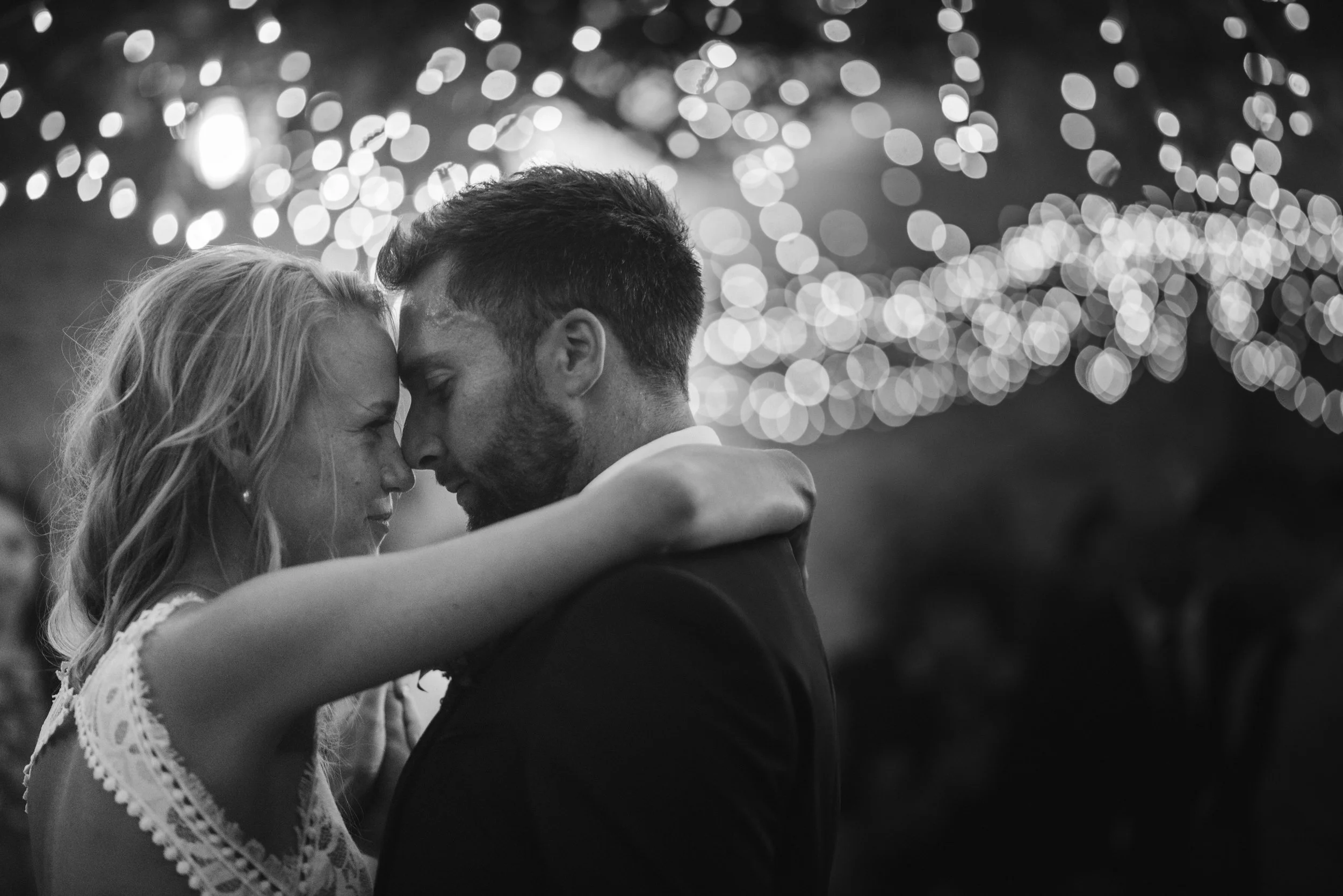 A black and white photo of a couple with their foreheads touching, arms around each other, in an intimate moment with blurry string lights in the background.