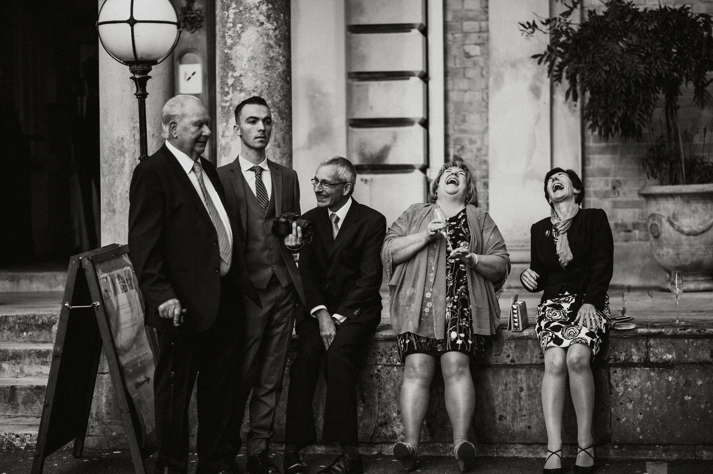 Group of people in formal attire laughing, standing and sitting outside a building, with a vintage lamp post and plants in the background, captured by award-winning Cornwall wedding photographer, Mark Shaw. 