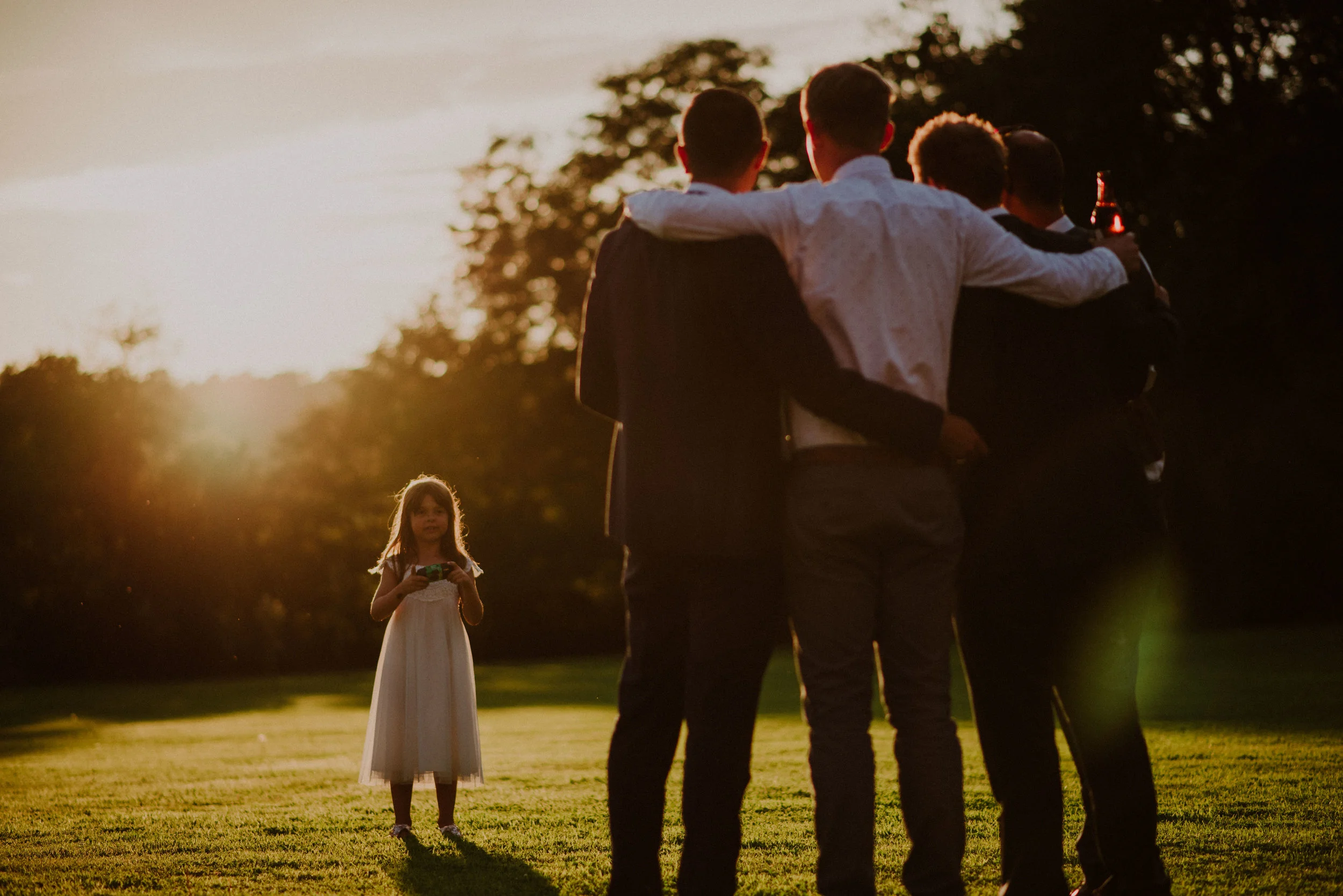 Little girl in white dress holding camera, taking photo of group of four men with arms around each other, standing on grass, at sunset.