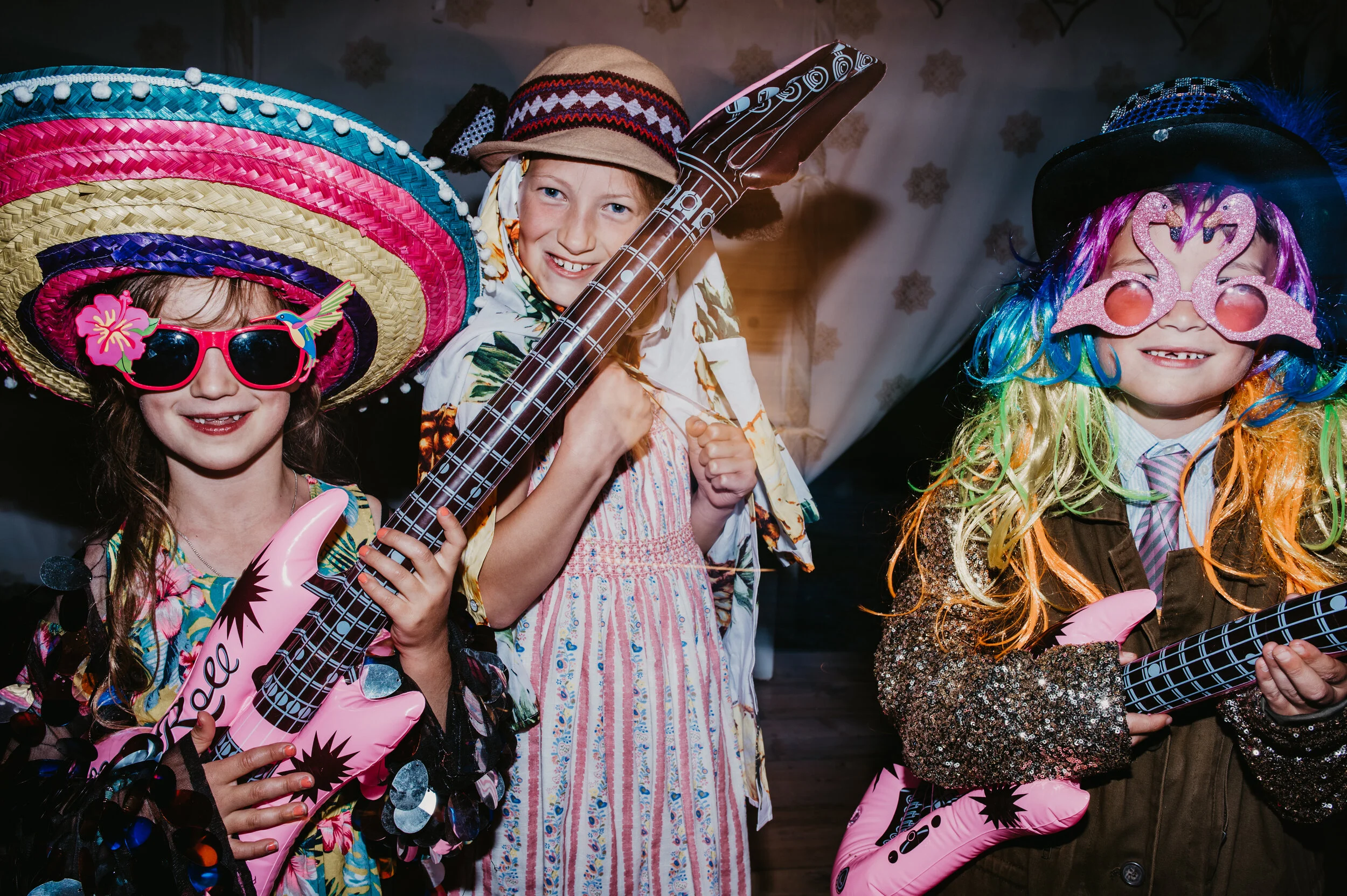 Three children dressed in colorful costumes with hats, sunglasses, and inflatable guitars, smiling and posing for a playful group photo, captured by award-winning Cornwall wedding photographer, Mark Shaw. 
