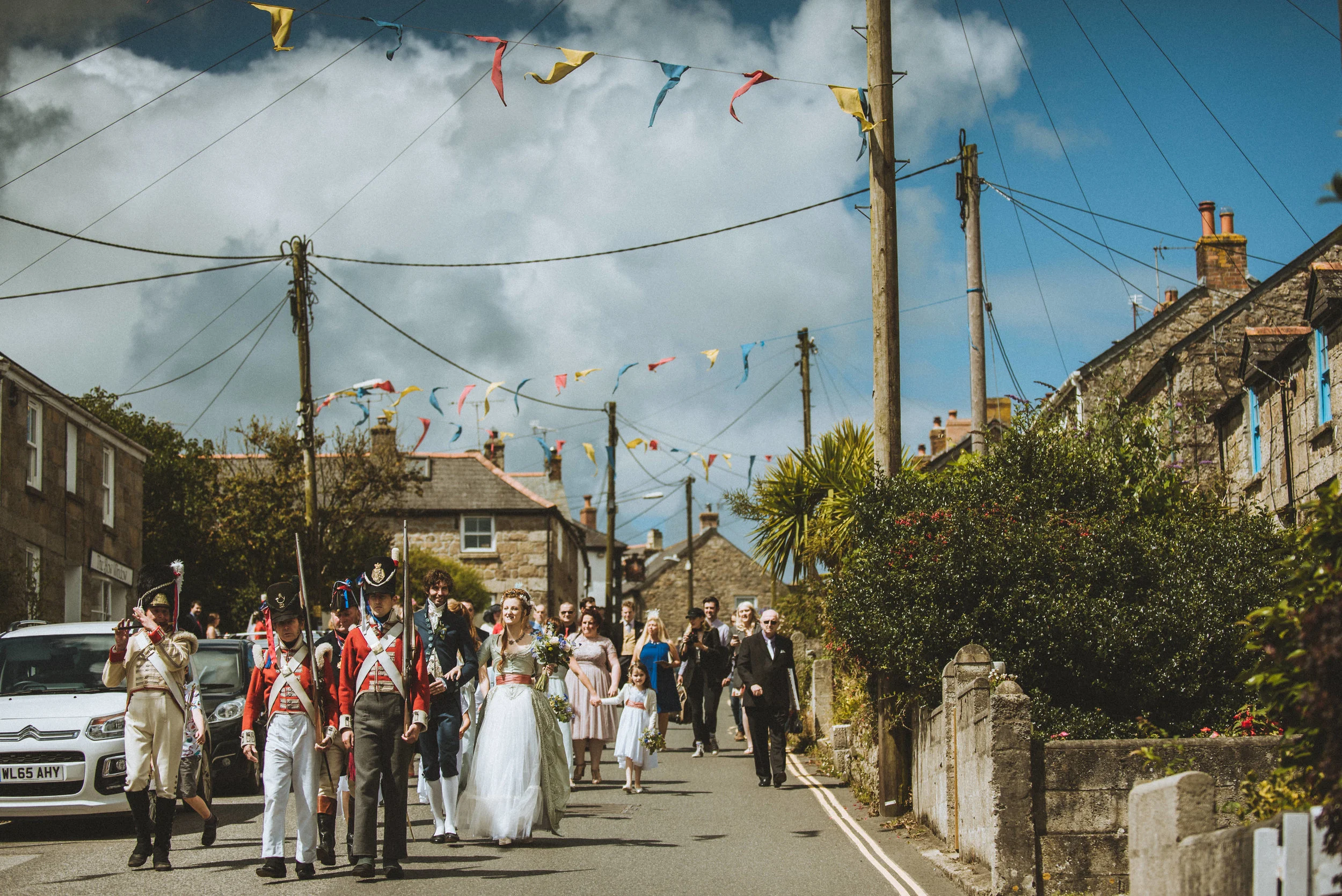 Wedding procession in a village street with people in historical military uniforms and traditional attire, captured by award-winning Cornwall wedding photographer, Mark Shaw. 