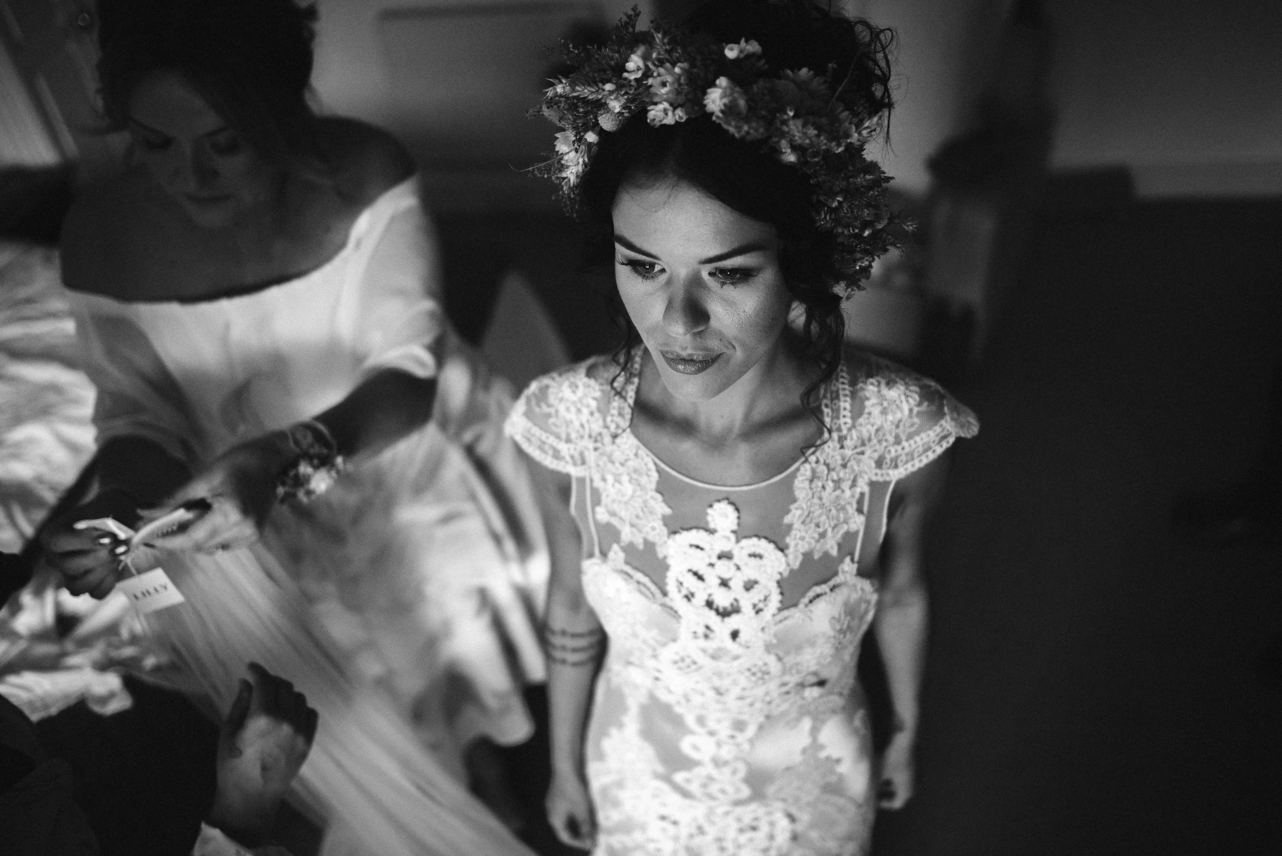 Black and white photo of a bride in a lace wedding dress with a floral crown, accompanied by another woman adjusting fabric.