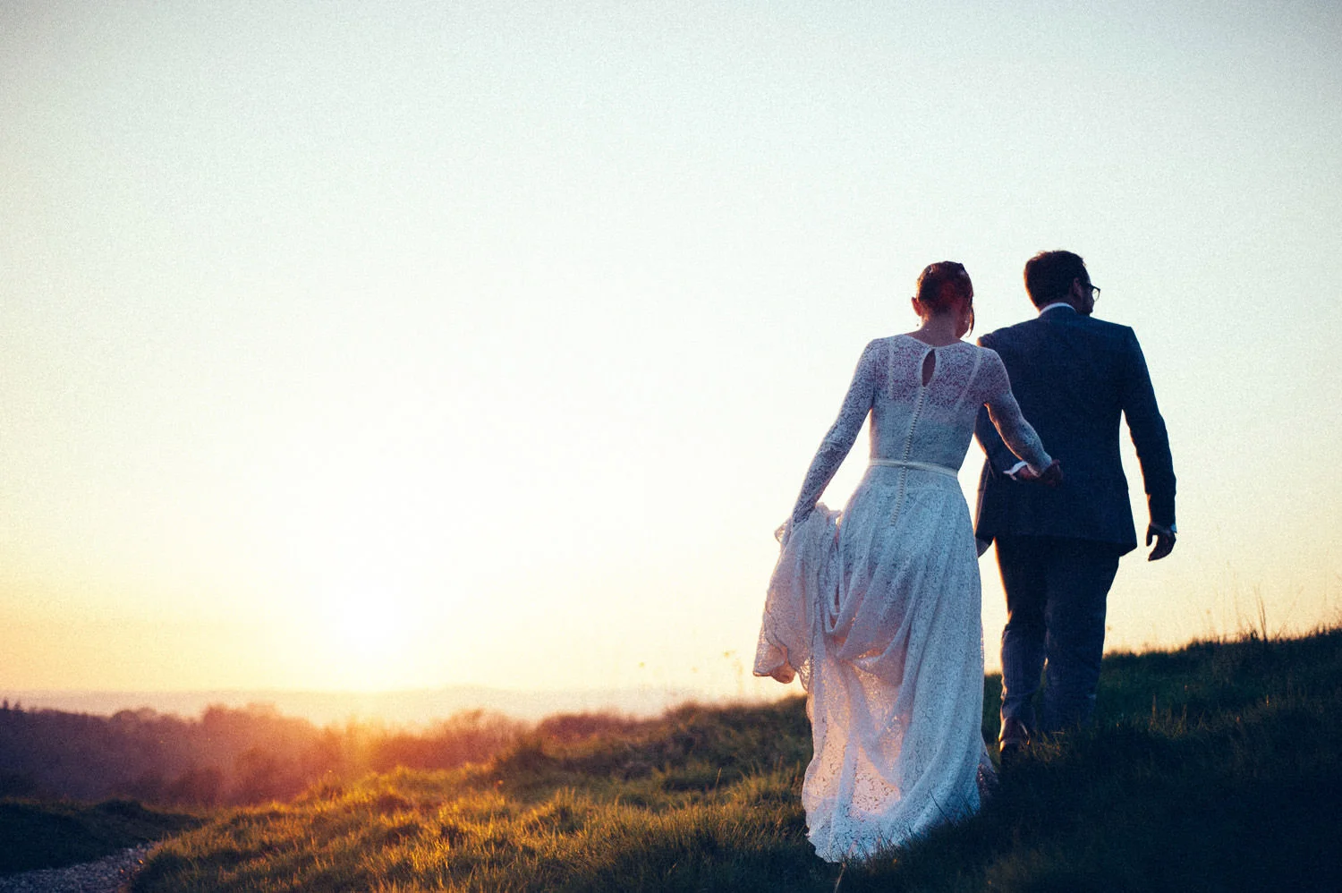 Bride and groom walking in a field at sunset, back view, captured by award-winning Cornwall wedding photographer, Mark Shaw. 