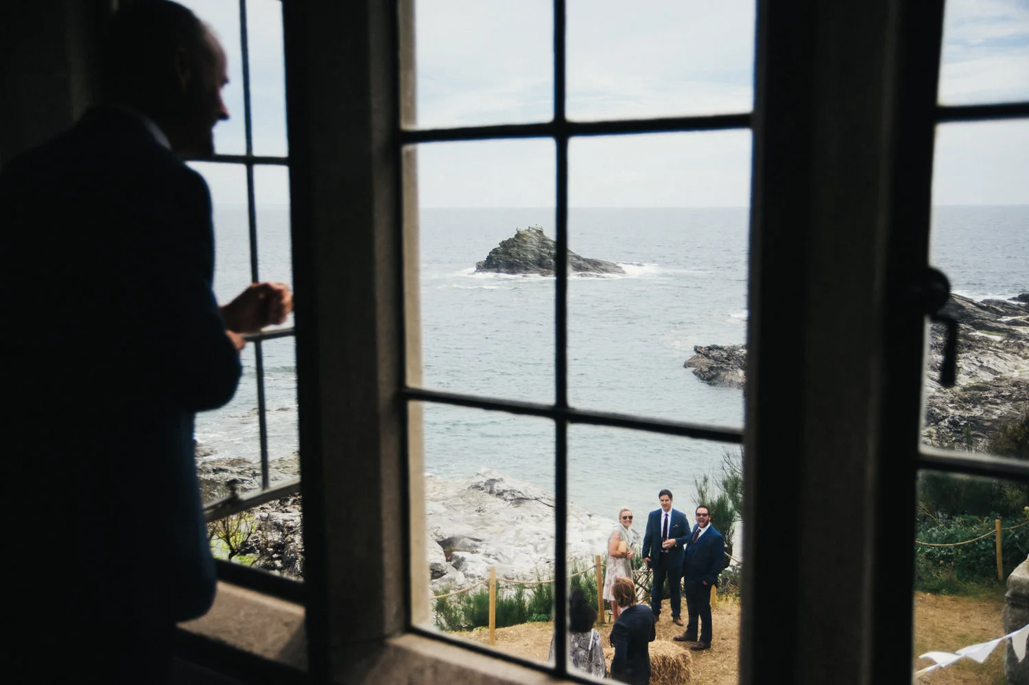 Group of wedding guests standing on a rocky coastal area viewed through a window as groom looks down to them, captured by award-winning Cornwall wedding photographer, Mark Shaw. 
