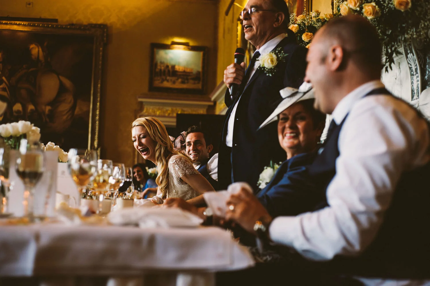 Wedding reception, guests laughing, man giving speech.