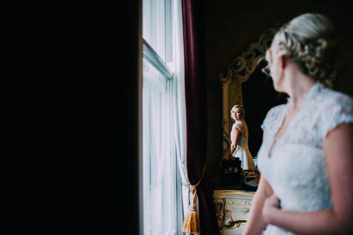 A bride in a wedding dress looking at her reflection in an ornate mirror, with her image visible in the mirror in front of her.