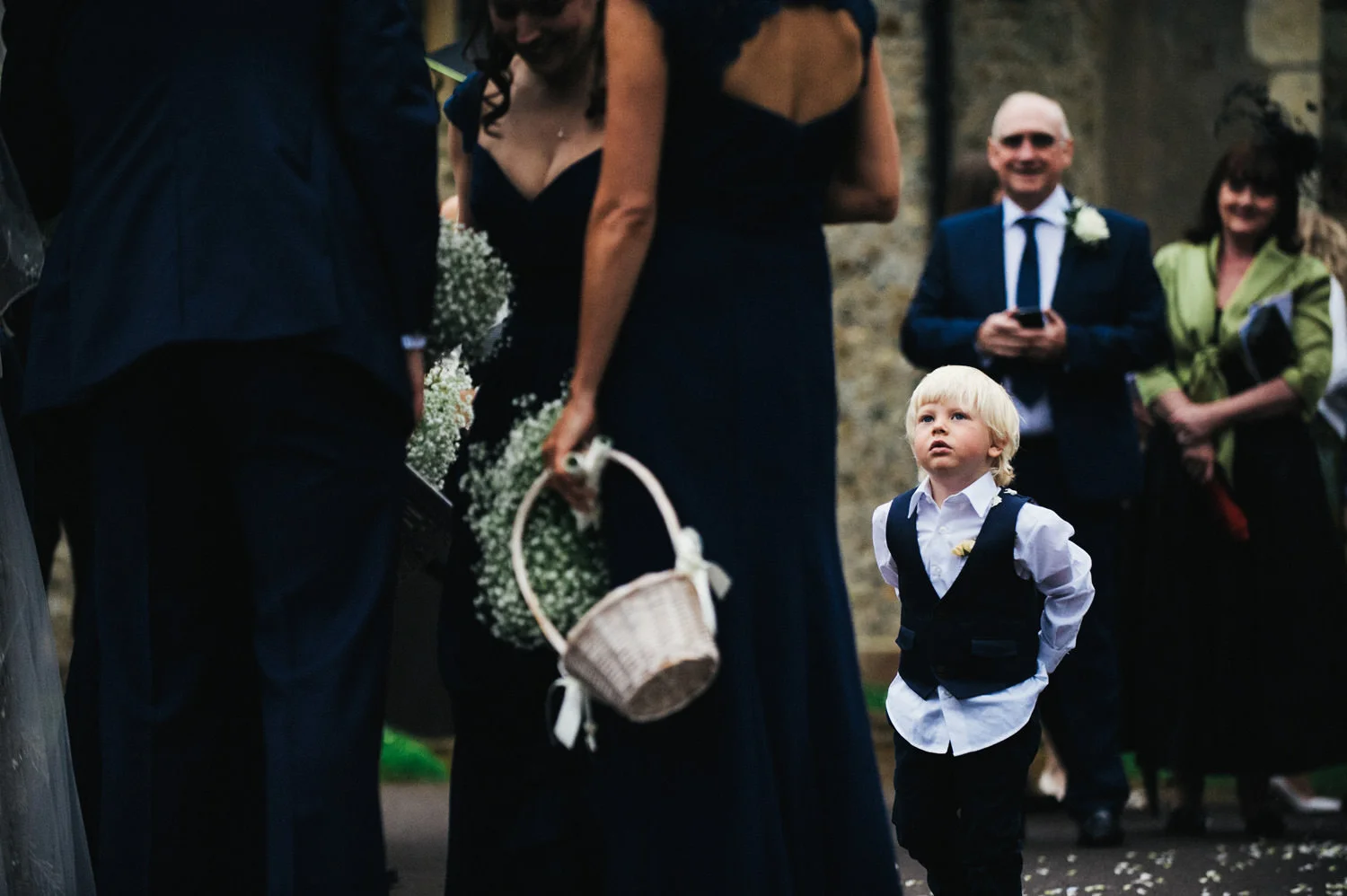 Young child in formal attire looking up at adults at a wedding ceremony, with people in dark clothing and a flower basket in the foreground, captured by award-winning Cornwall wedding photographer, Mark Shaw. 