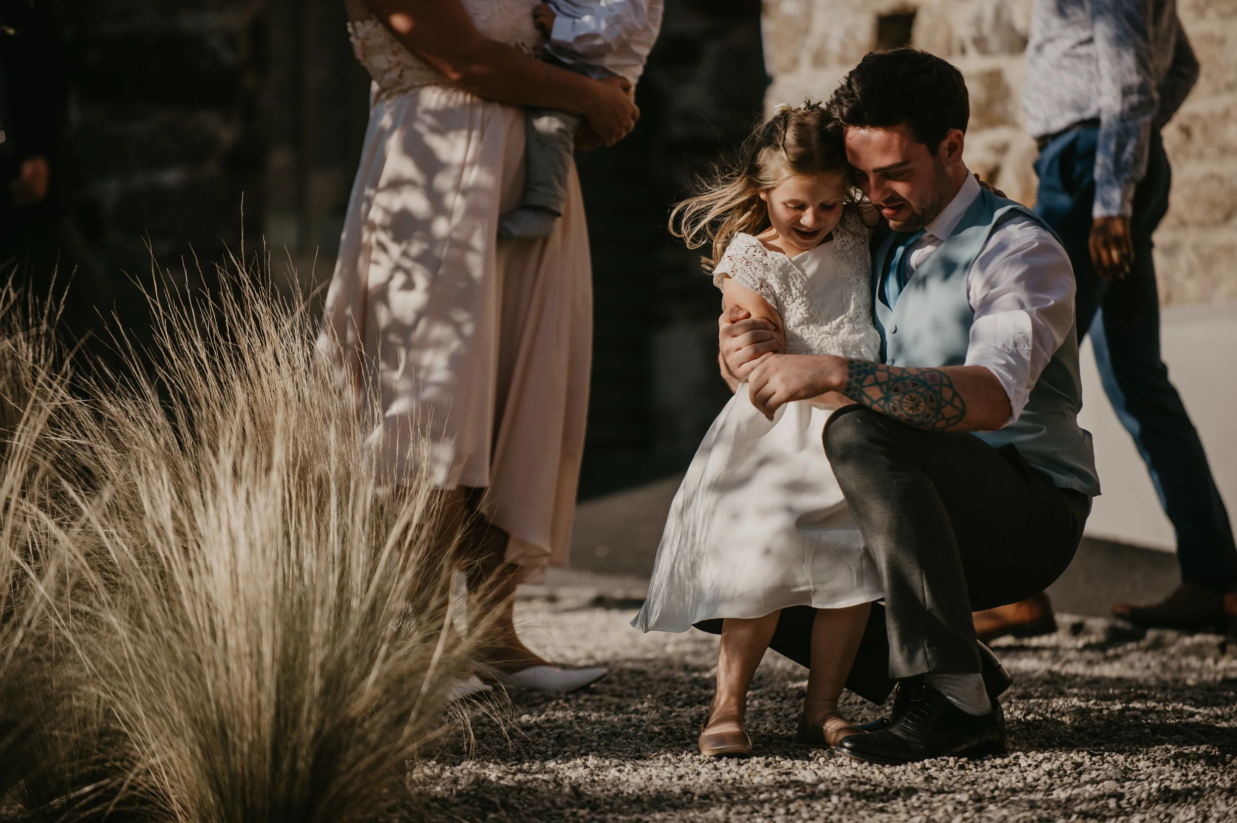 A man crouching, holding a young girl in a white dress, standing outdoors in a sunlit garden, captured by award-winning Cornwall wedding photographer, Mark Shaw. 