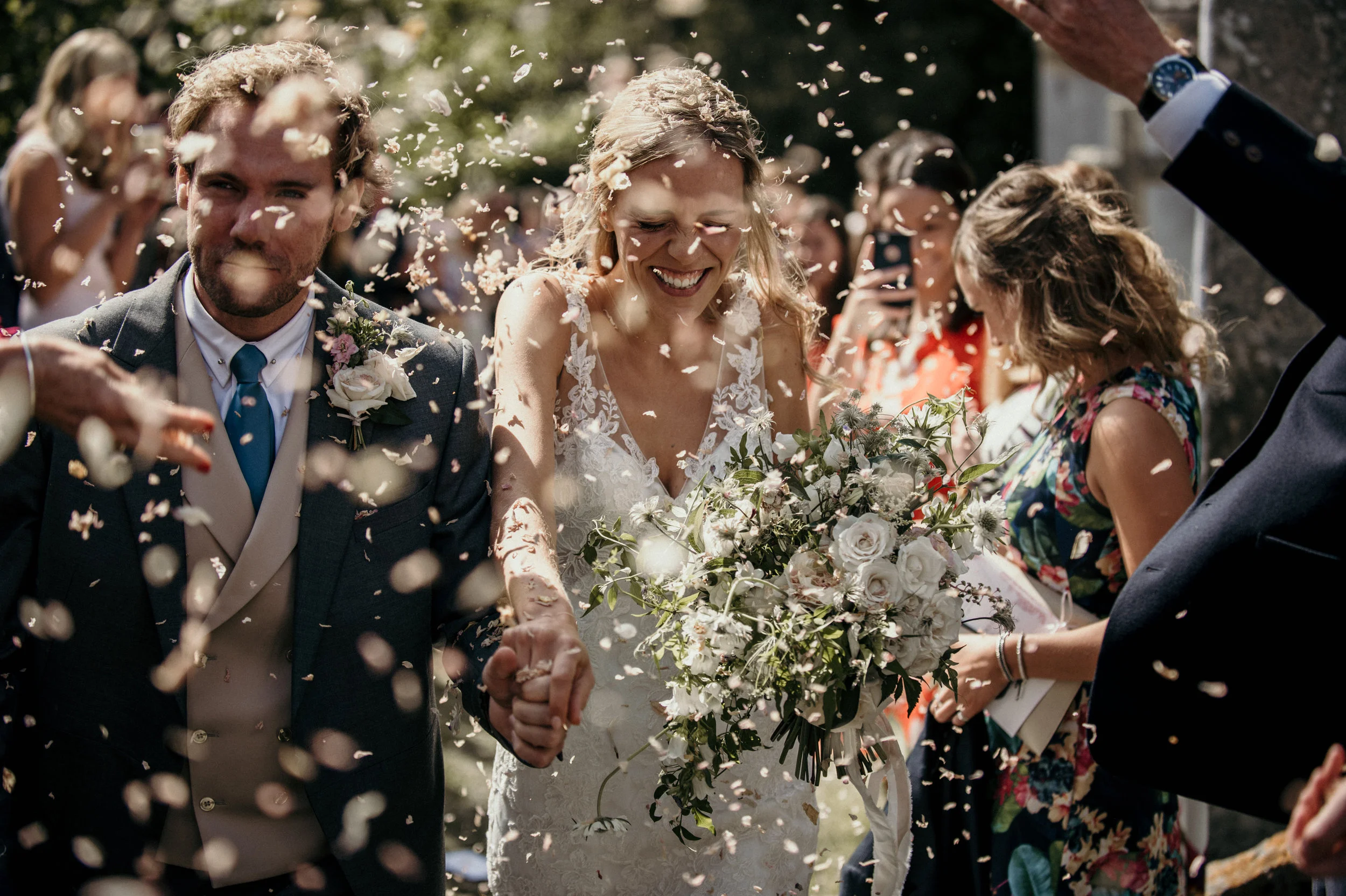 Bride and groom exiting wedding venue, smiling, with flower confetti, bride holding bouquet, guests in background.