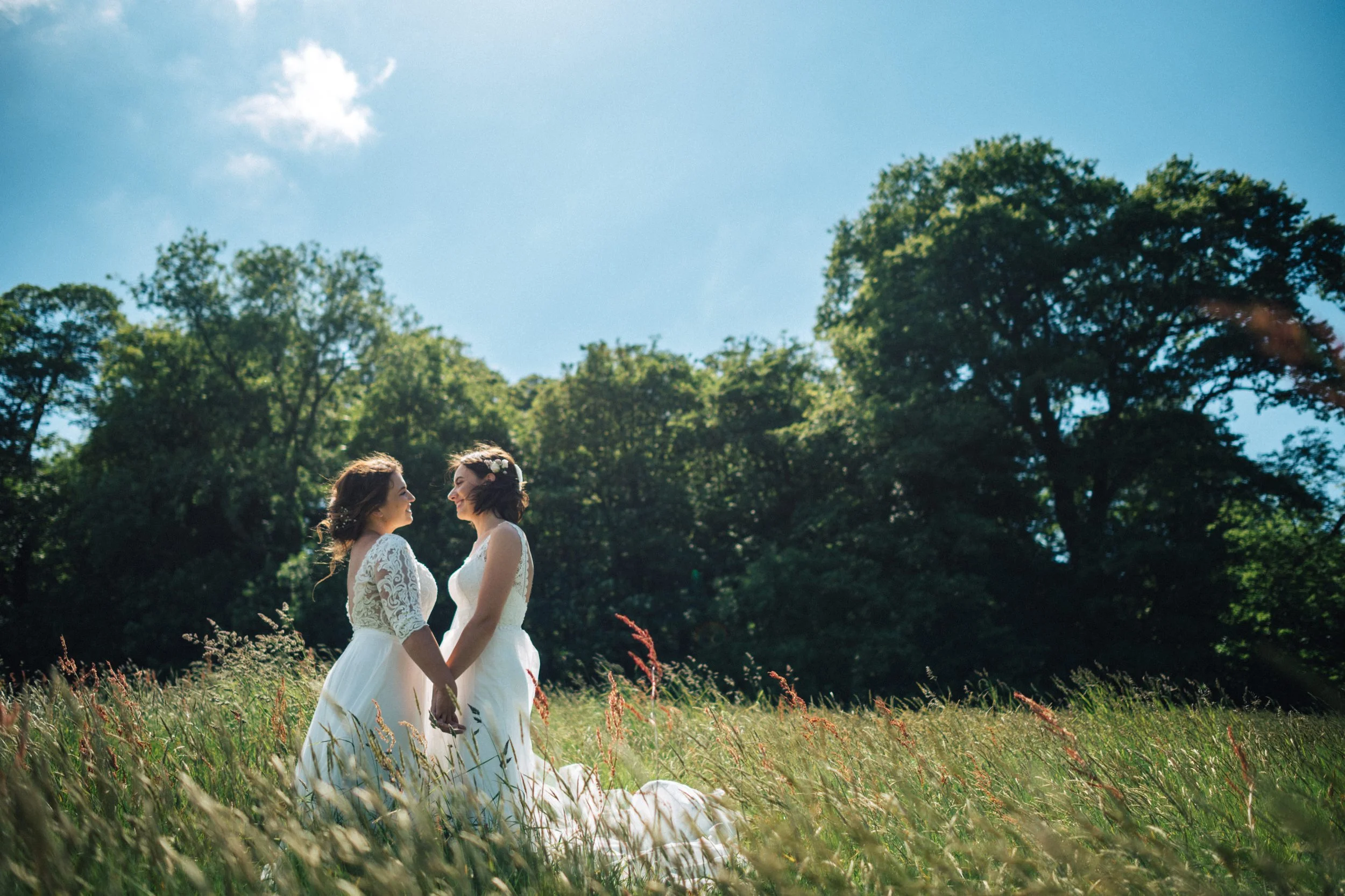 Two women in wedding dresses holding hands and smiling at each other in a grassy field with trees and a bright blue sky in the background.