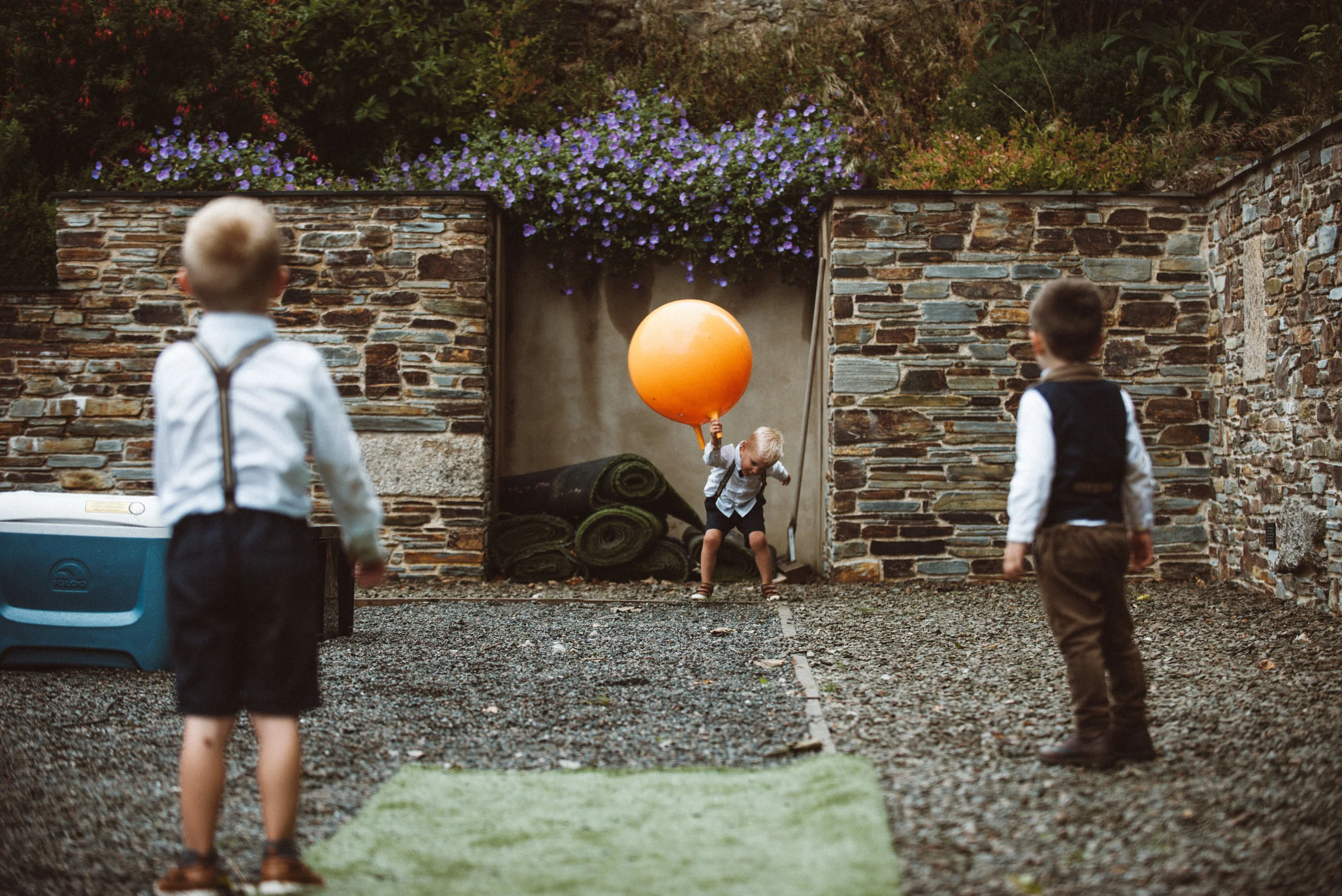 Three young boys play in a garden with stone walls and purple flowers, one holding a large orange balloon.