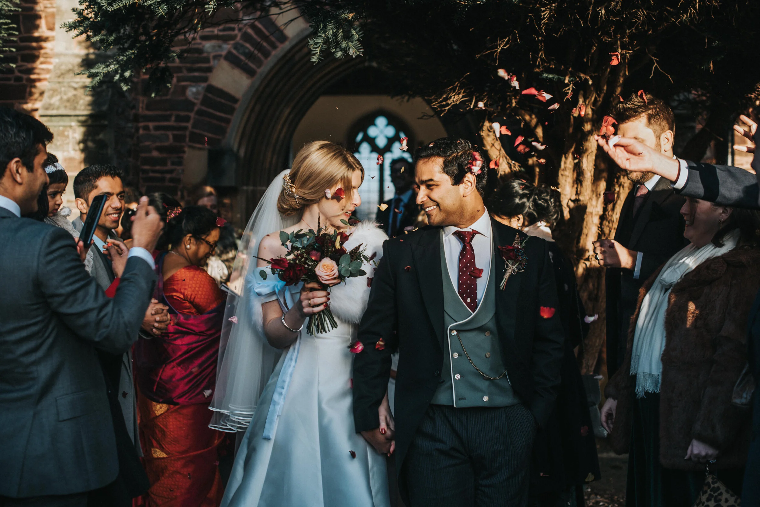 Bride and groom exiting a church, holding hands, surrounded by guests, with flower petals in the air, captured by award-winning Cornwall wedding photographer, Mark Shaw. 