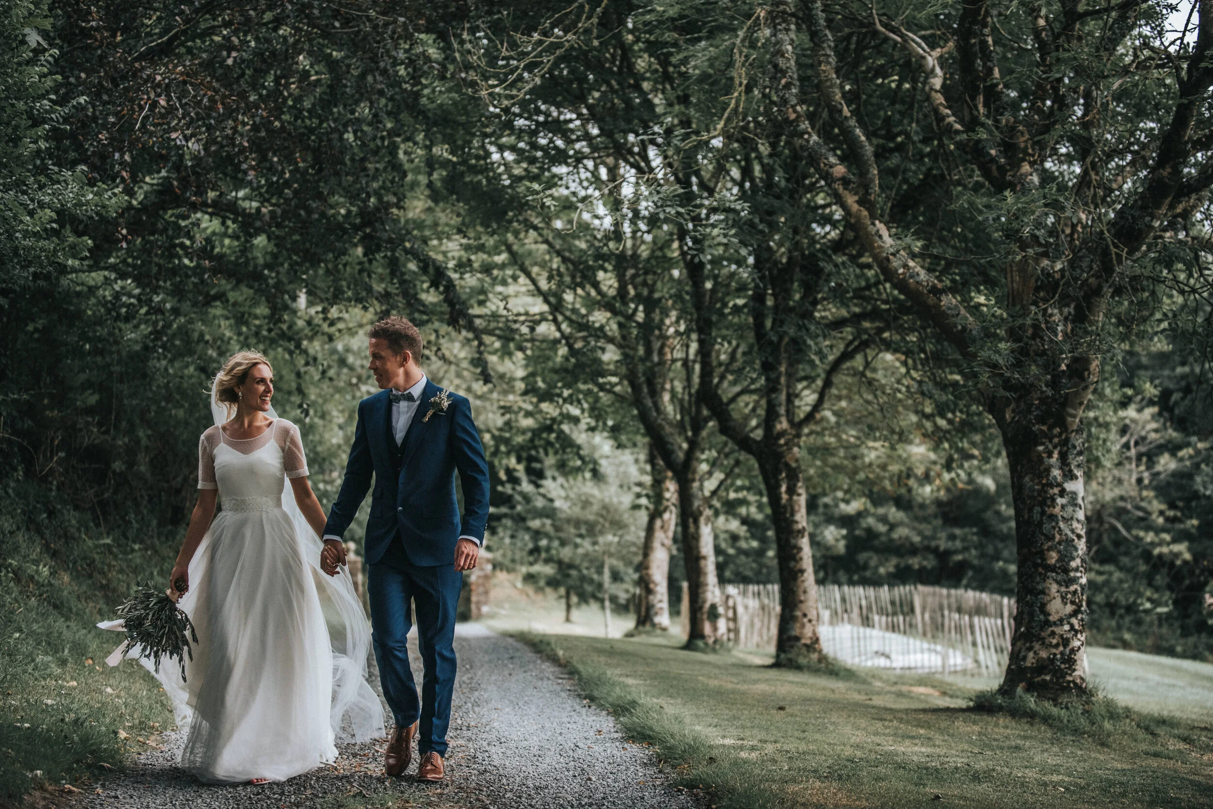 Bride and groom walking hand in hand on a tree-lined path, captured by award-winning Cornwall wedding photographer, Mark Shaw. 