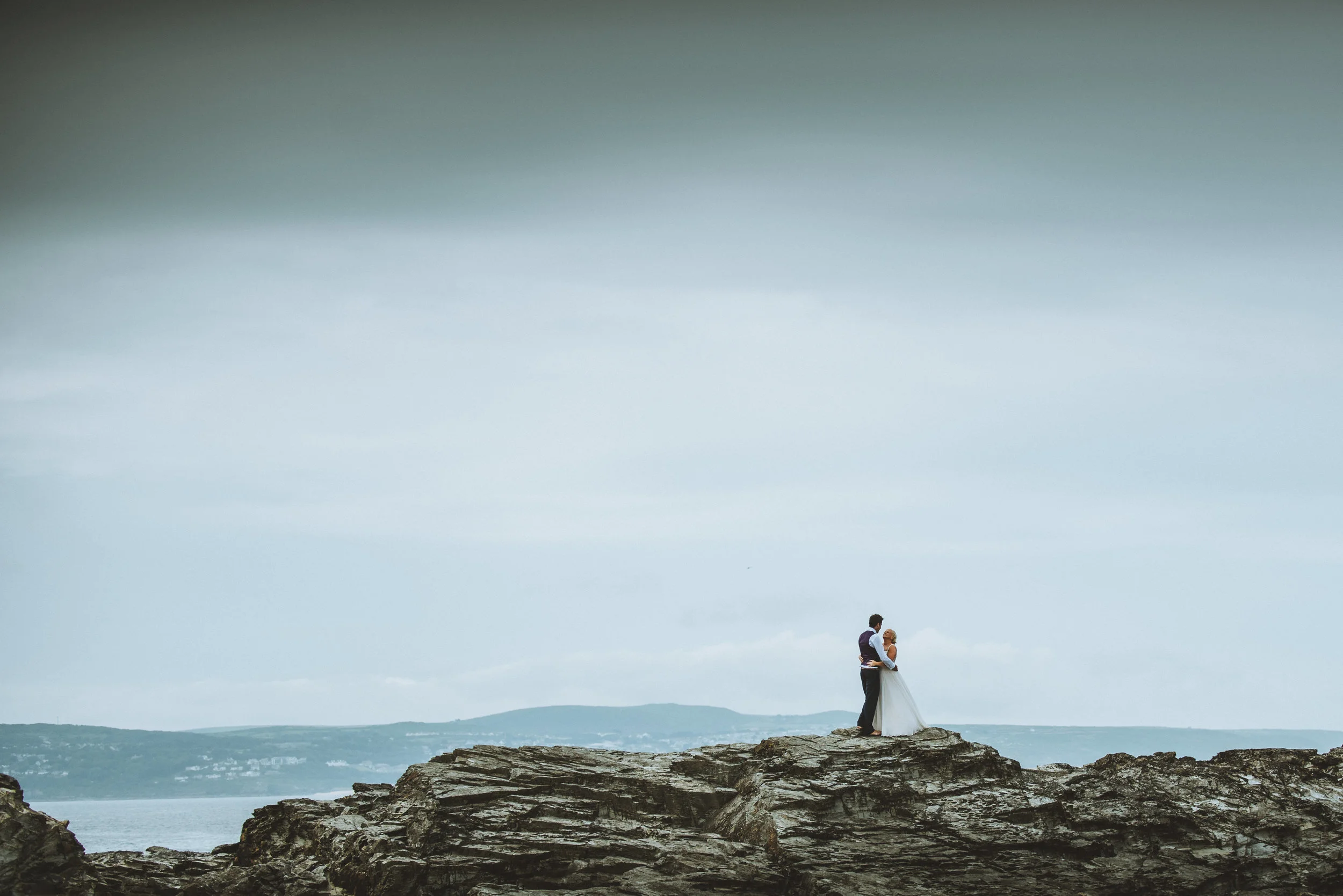 A couple in wedding attire standing on rocky terrain near water, with overcast skies in the background.