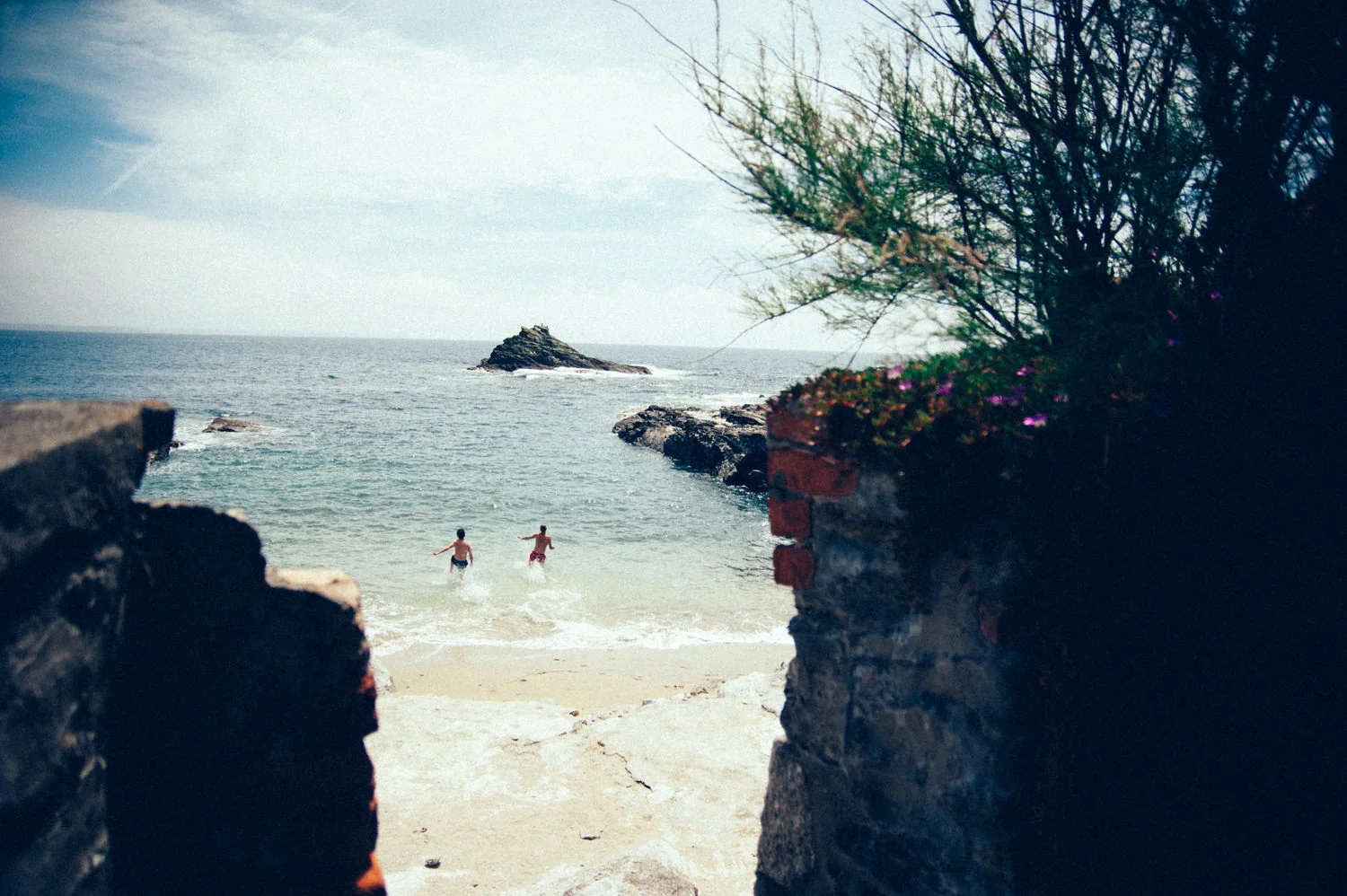 View of a beach with two children playing in the water, framed by brick walls and greenery, ocean waves, rocky outcrop, and cloudy sky in the background.