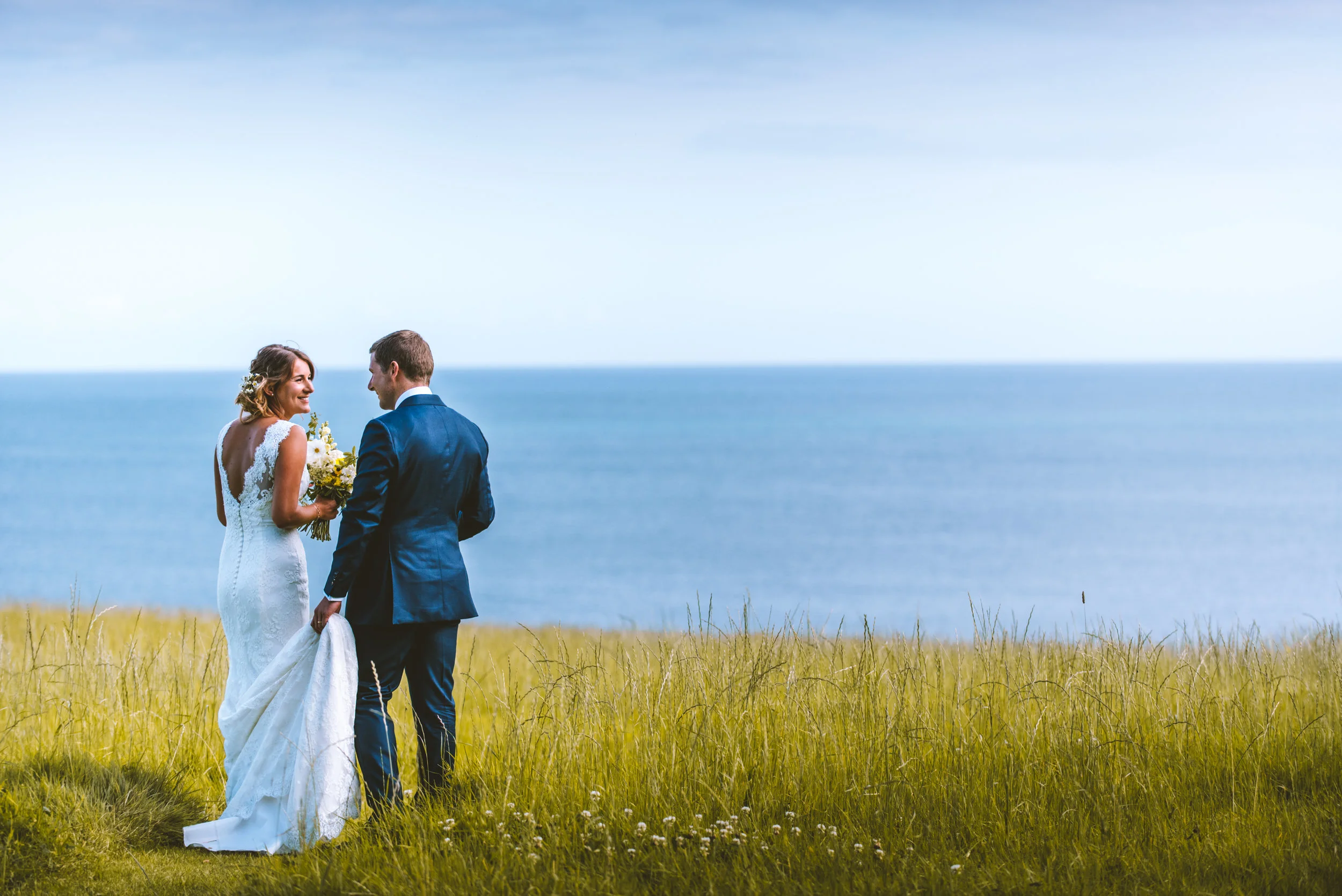 A couple dressed in wedding attire standing in a grassy field near the ocean, facing each other and holding a bouquet of flowers.