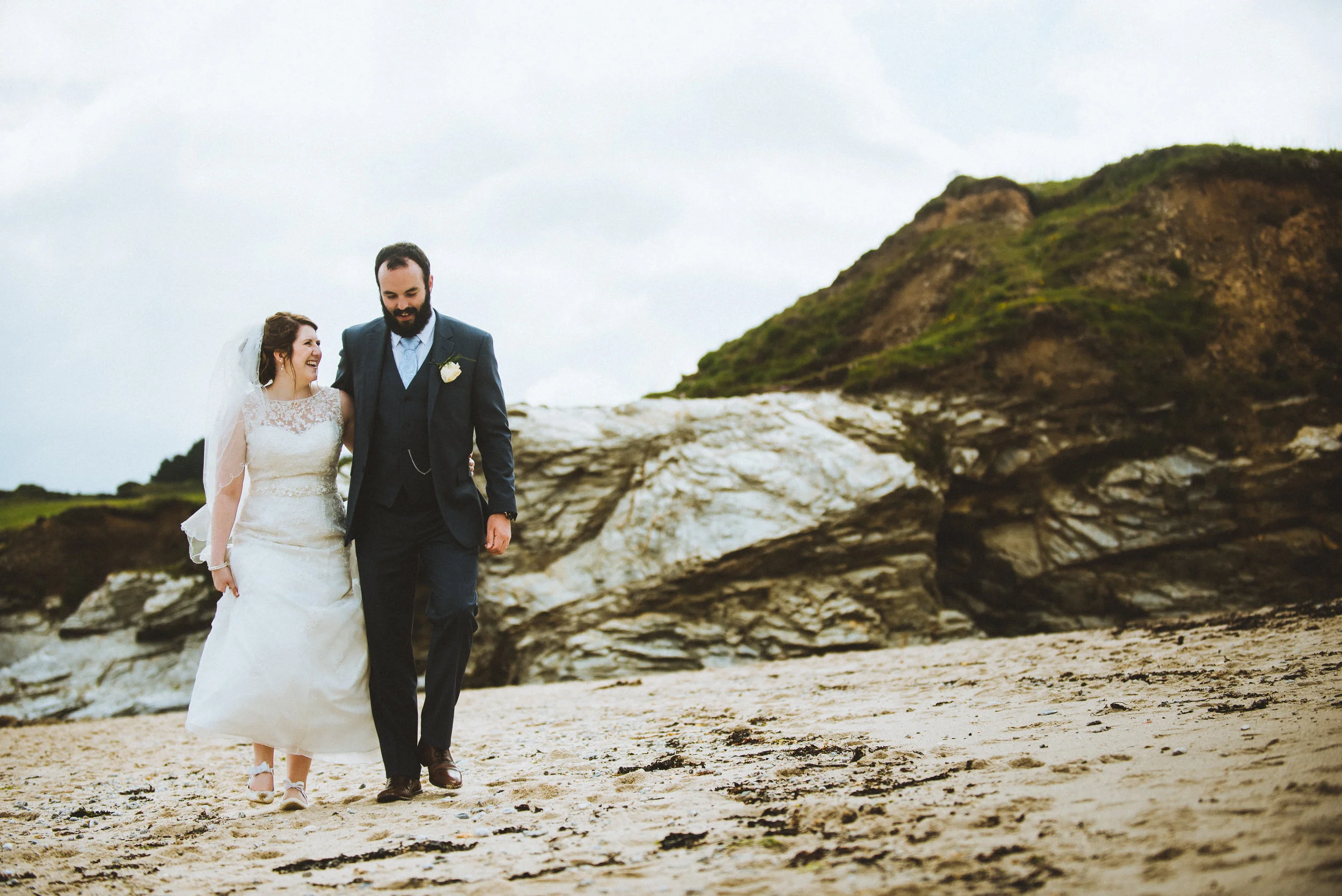 A bride and groom walking together on a sandy beach, smiling and laughing, with rocky formations and a grassy hill in the background.