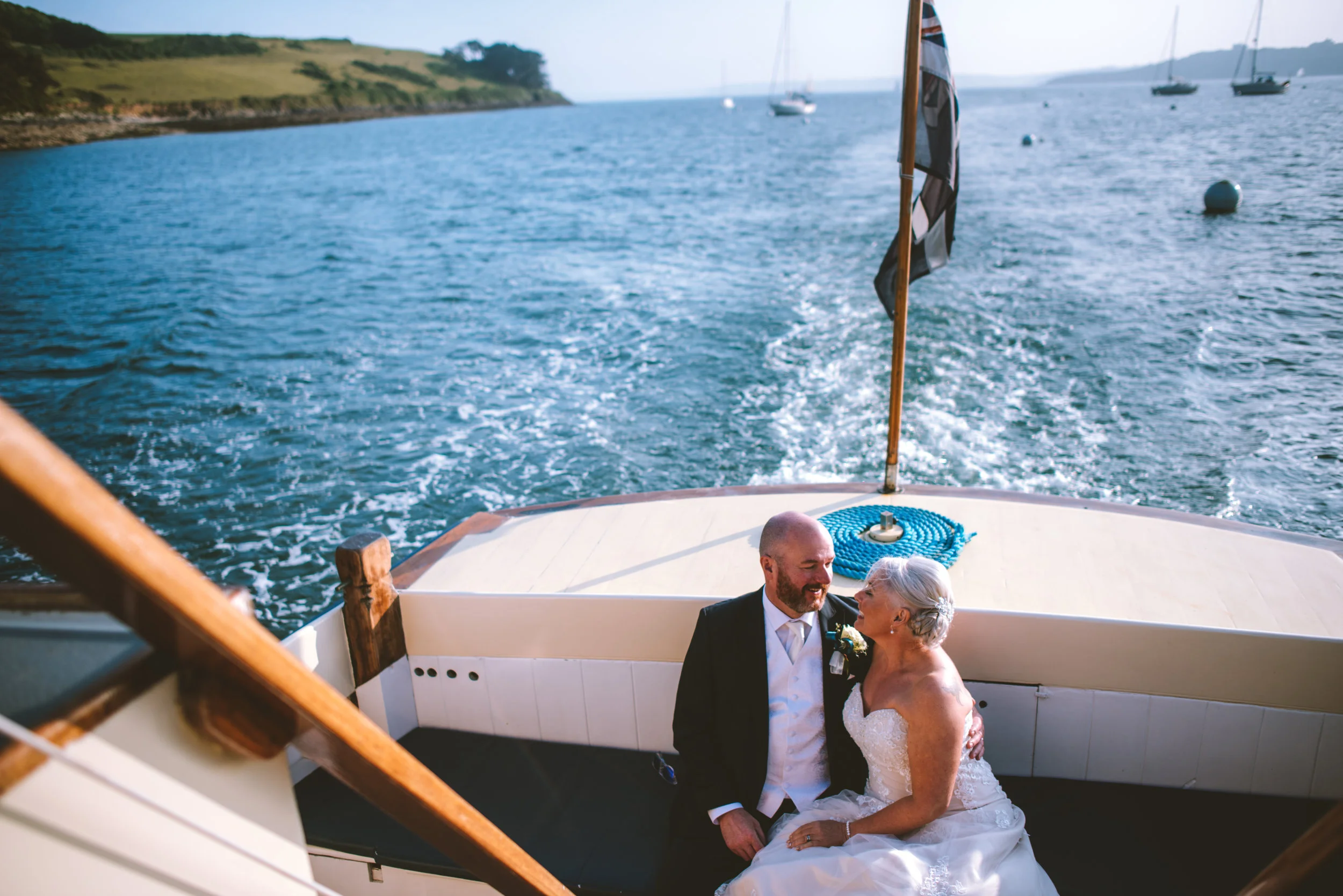 A newlywed couple in wedding attire sitting on the deck of a boat during daytime, with water and boats visible in the background.