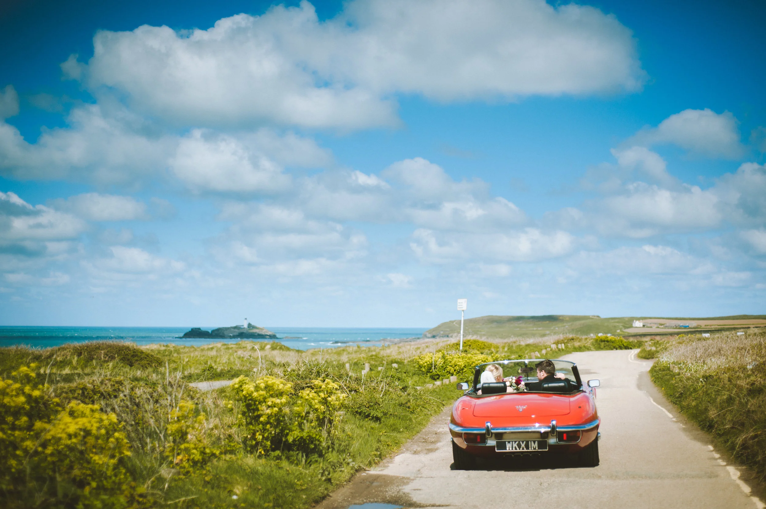 Bride and groom in convertible car driving along Gwithian coastal road captured by award-winning Cornwall wedding photographer Mark Shaw