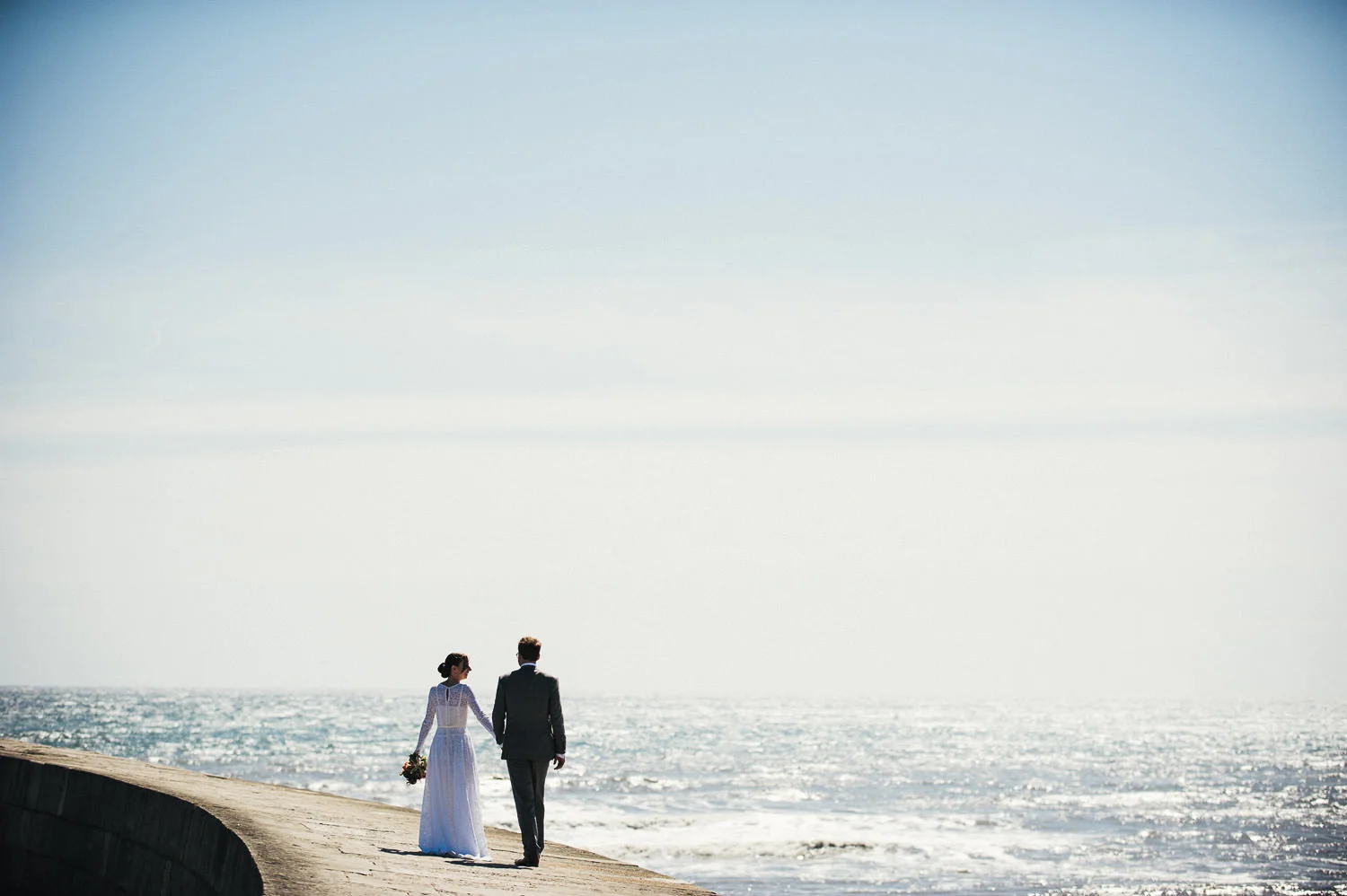 A bride and groom walking hand in hand along a seaside promenade during daytime, with the ocean and sky in the background.