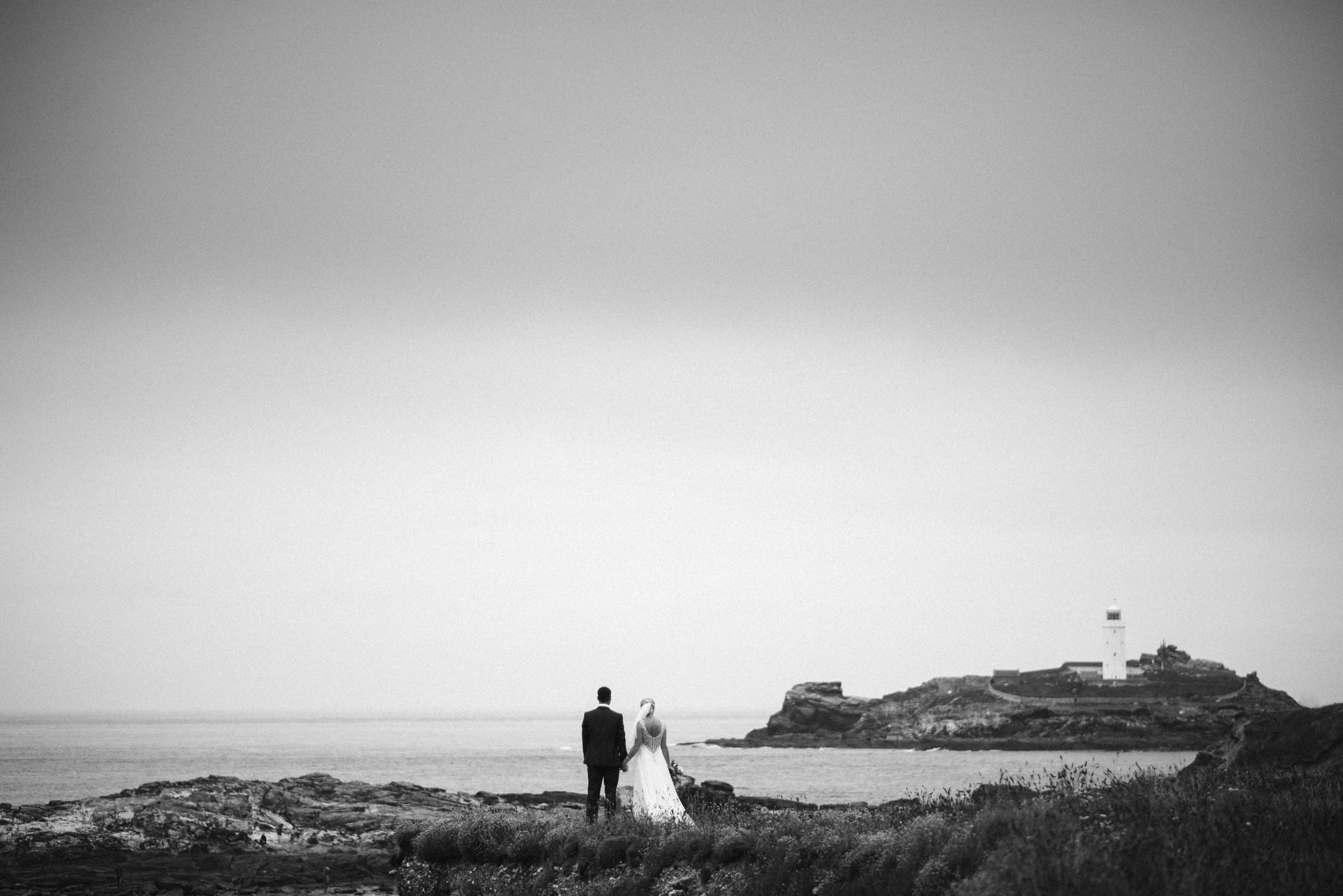 Black and white photo of a bride and groom standing on a rocky shoreline, holding hands and facing a distant lighthouse on a small island, with water and sky in the background.