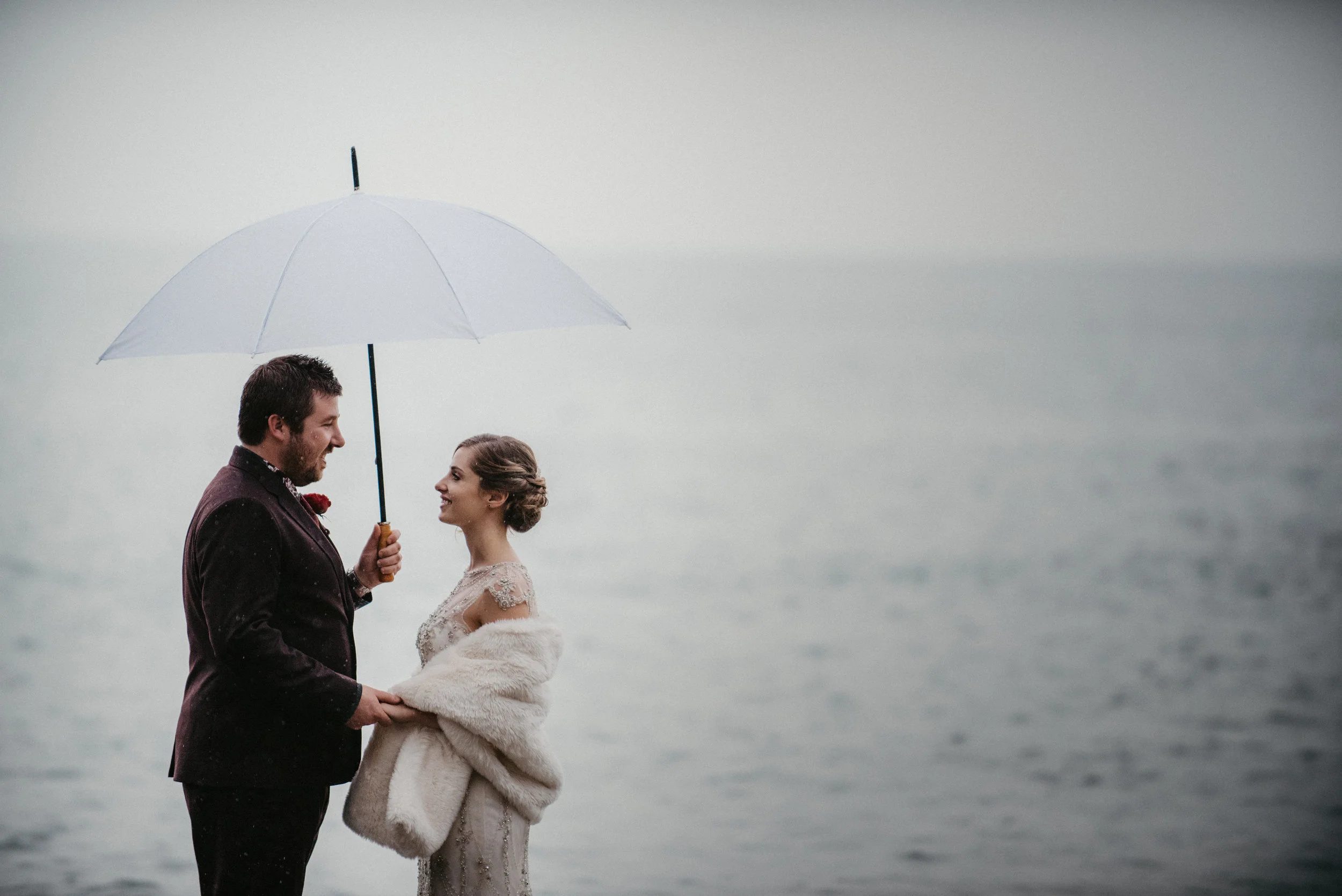 A bride and groom holding hands under an umbrella by the water, smiling at each other.