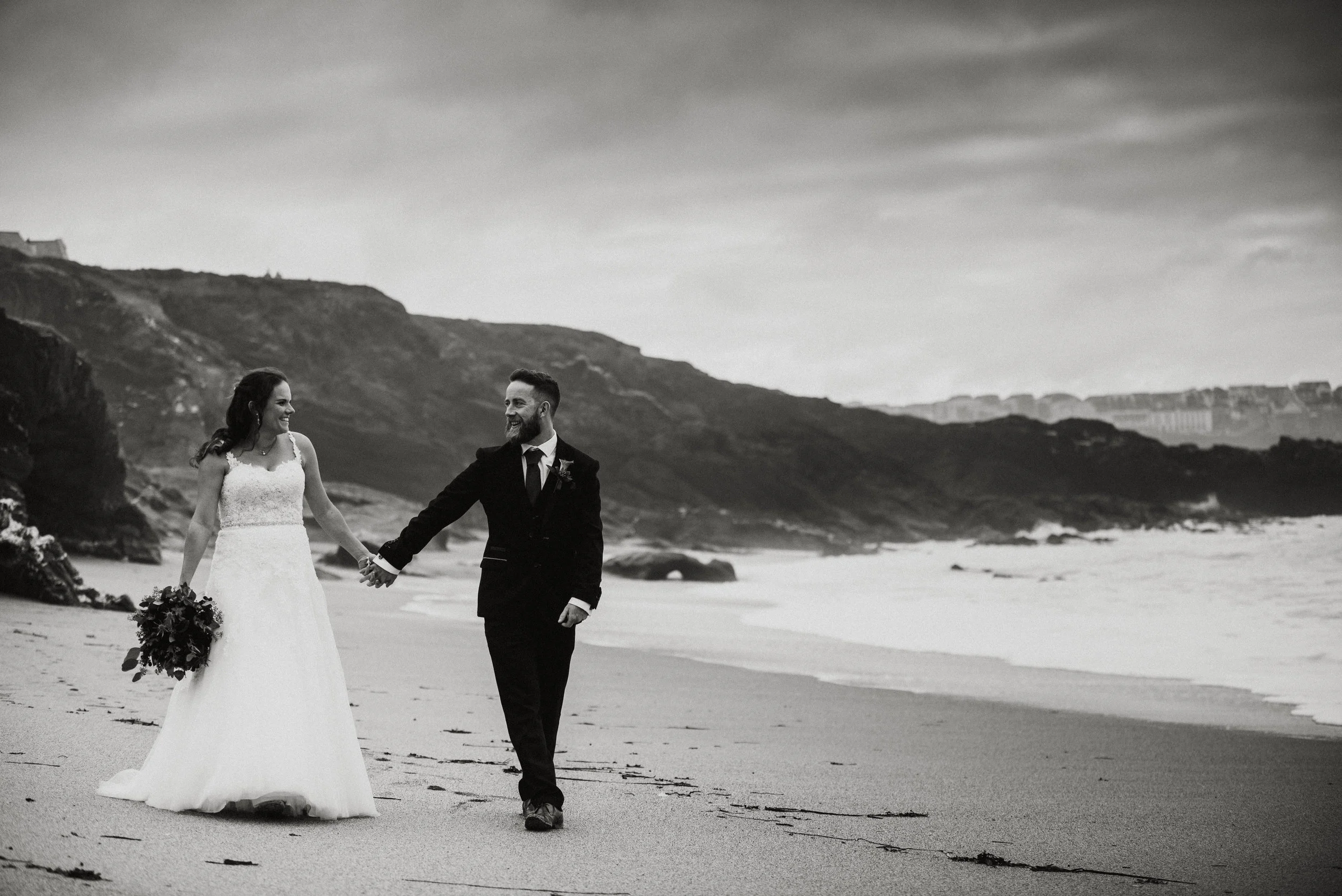 Black and white photo of a bride and groom walking hand in hand on a beach, with cliffs and ocean in the background.