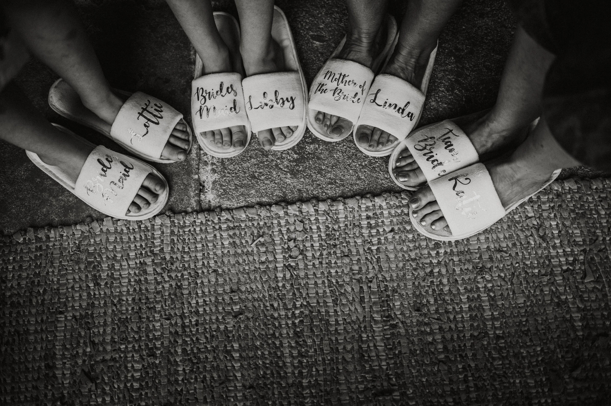 Six people wearing matching slippers with handwritten names and roles, standing in a circle on a textured ground, all viewed from above.
