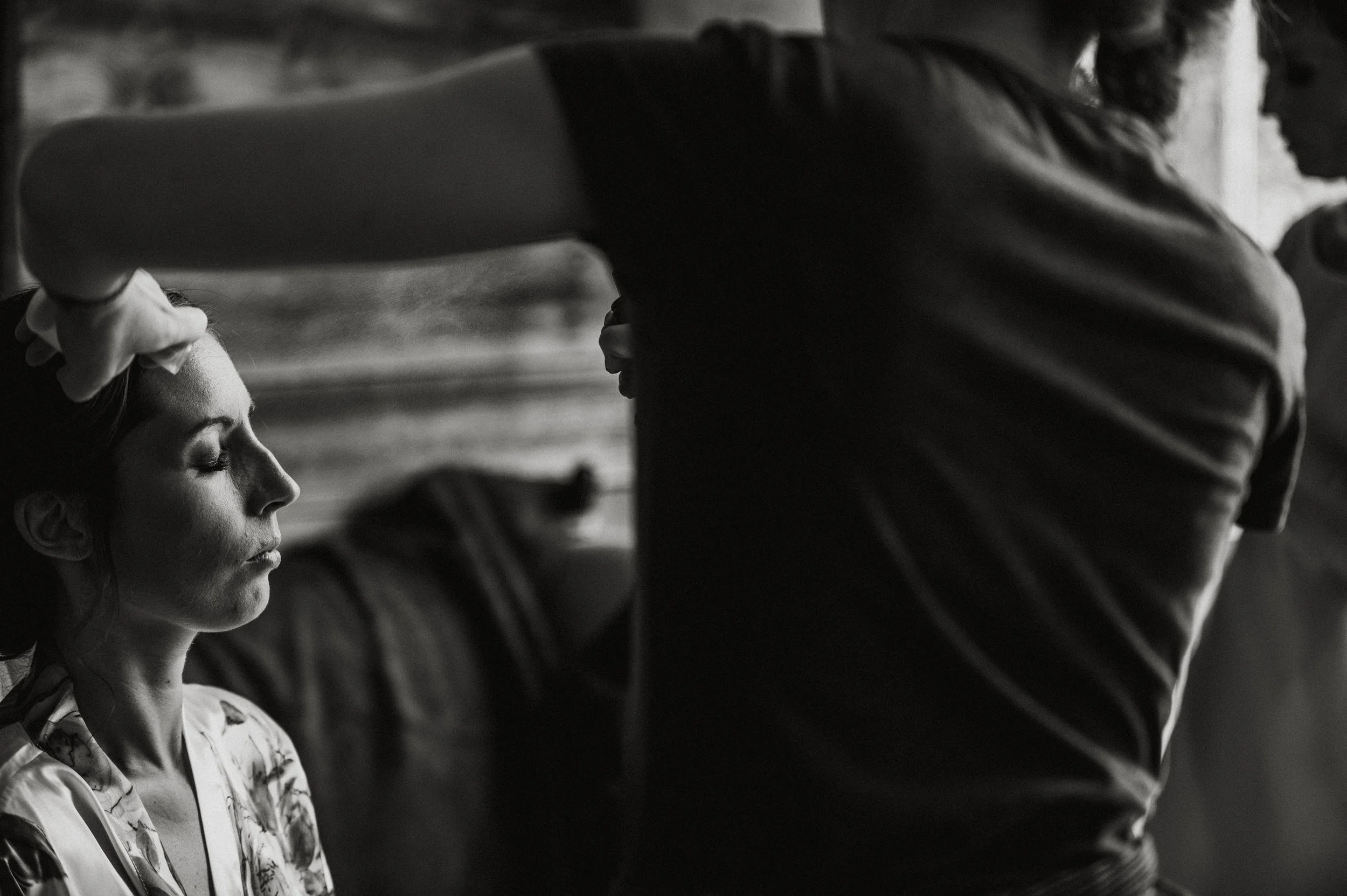 A woman with closed eyes and relaxed expression, having her makeup or hair done by a person who is partially visible, wearing a dark shirt.