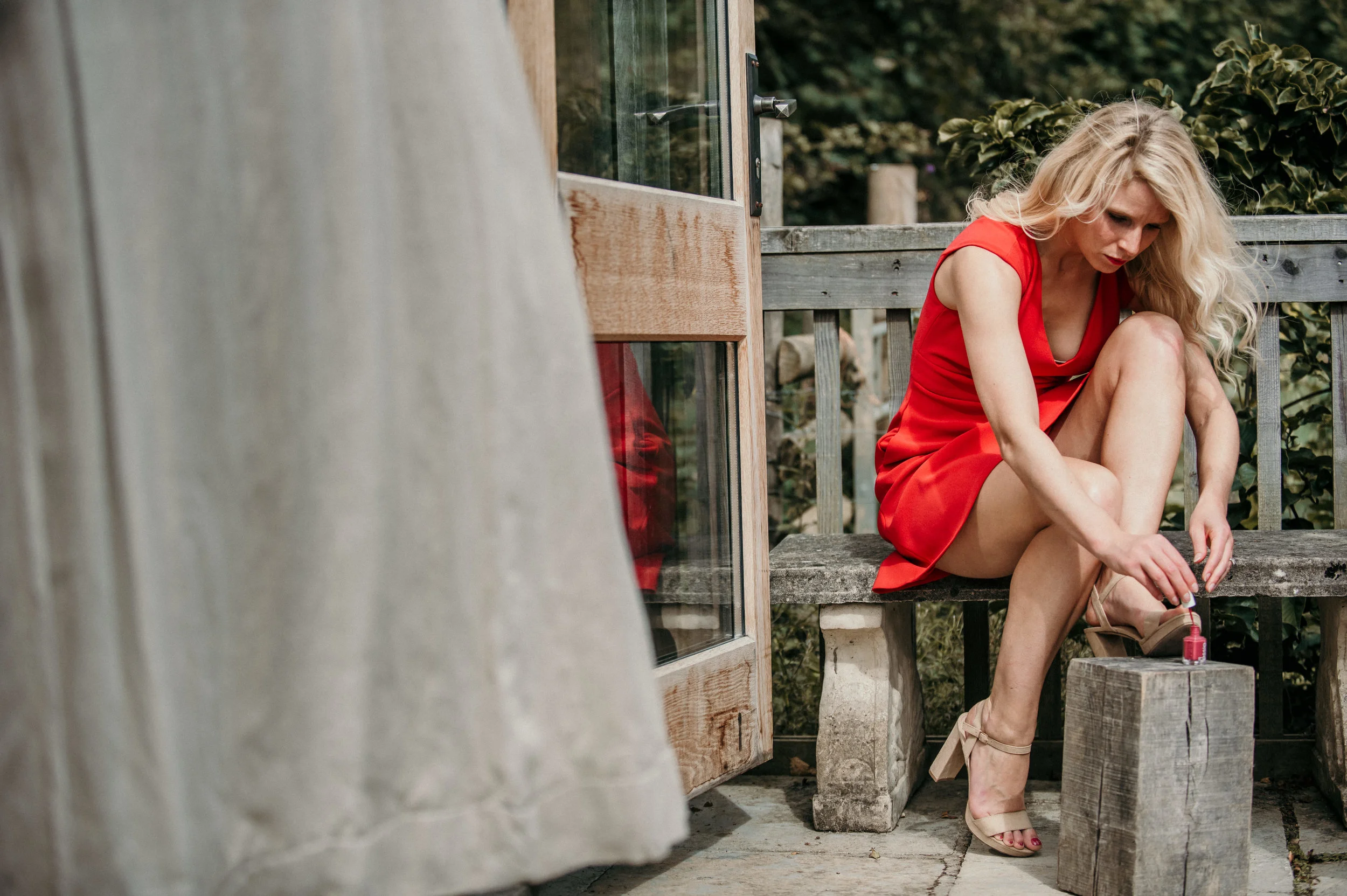 Woman in a red dress sitting on a wooden bench outside, applying pink nail polish to her toenails.