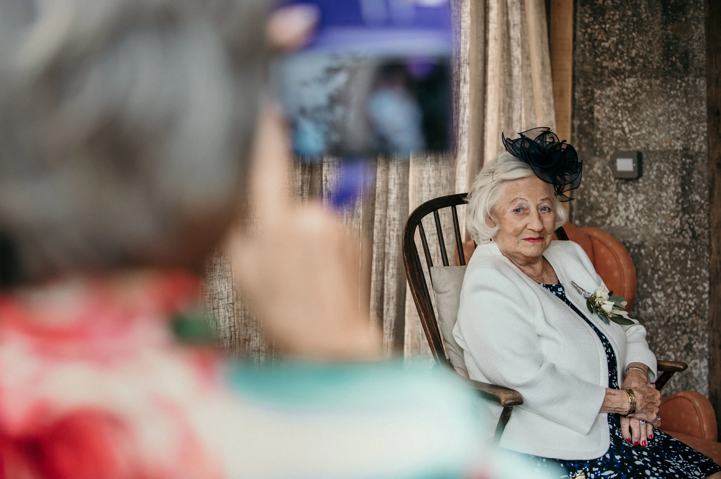 An elderly woman with white hair wearing a white blazer and a black floral dress with a black fascinator hat sitting on a chair at a social event, looking at the camera. Other guests are out of focus in the foreground.