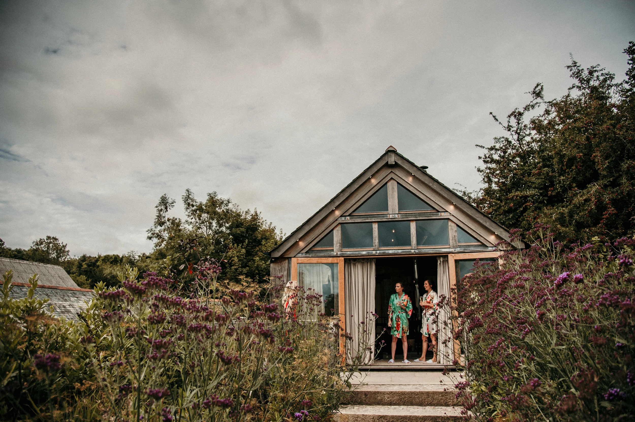 Two women standing in the doorway of a glass house, surrounded by purple flowers and greenery, during daytime with cloudy sky.