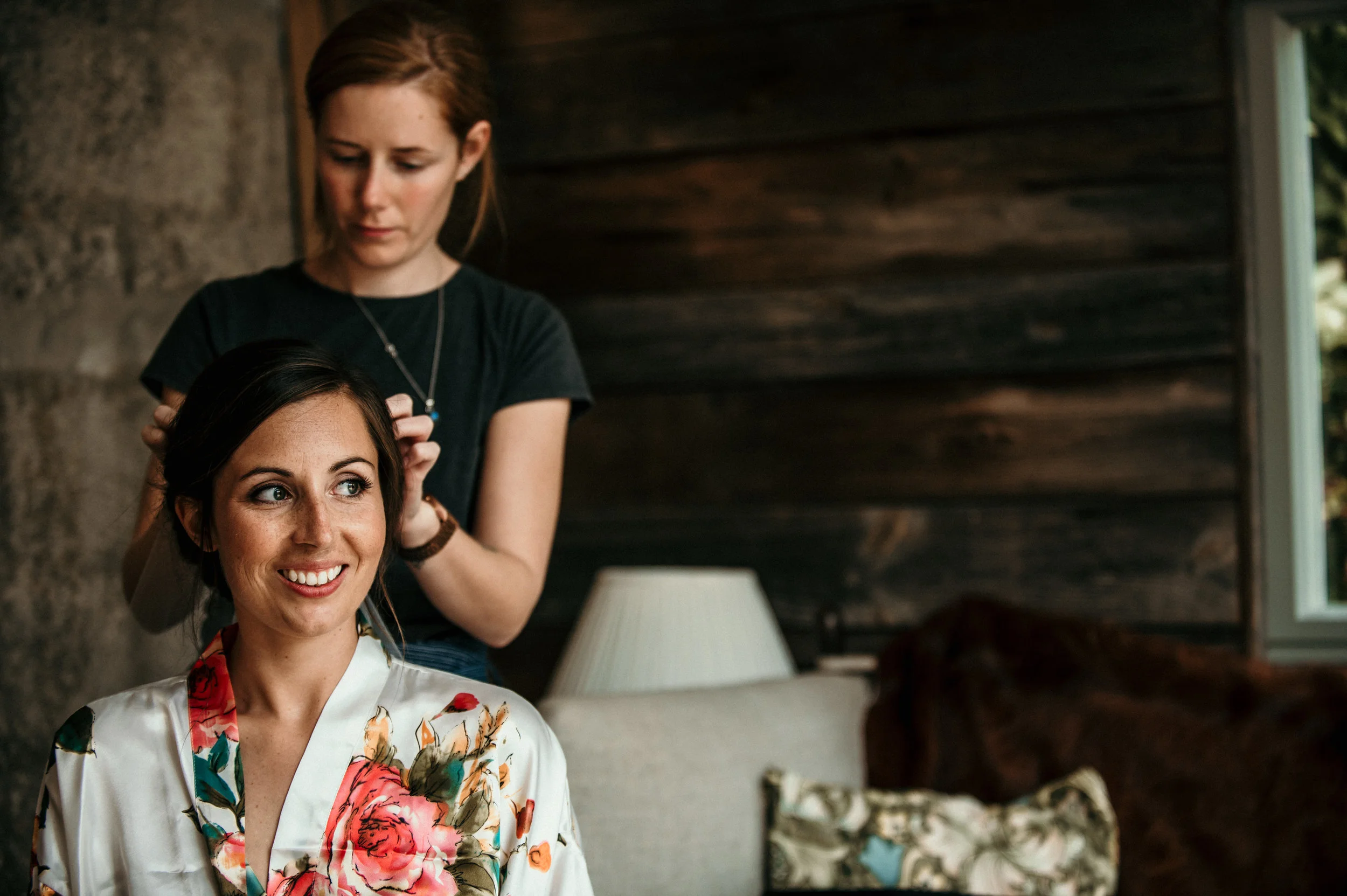 A woman with dark hair getting her hair styled by a hairdresser in a cozy room with wooden walls and a window.