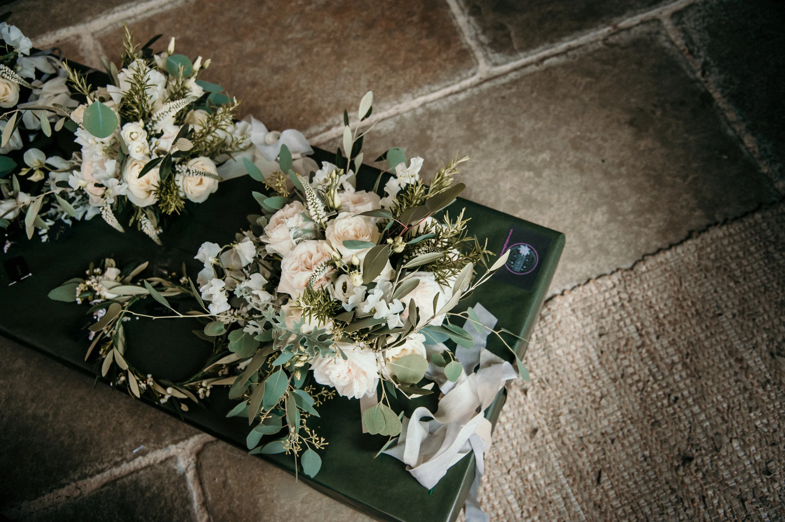 White and green floral wreath on a dark green bench near a brick wall and beige mat.