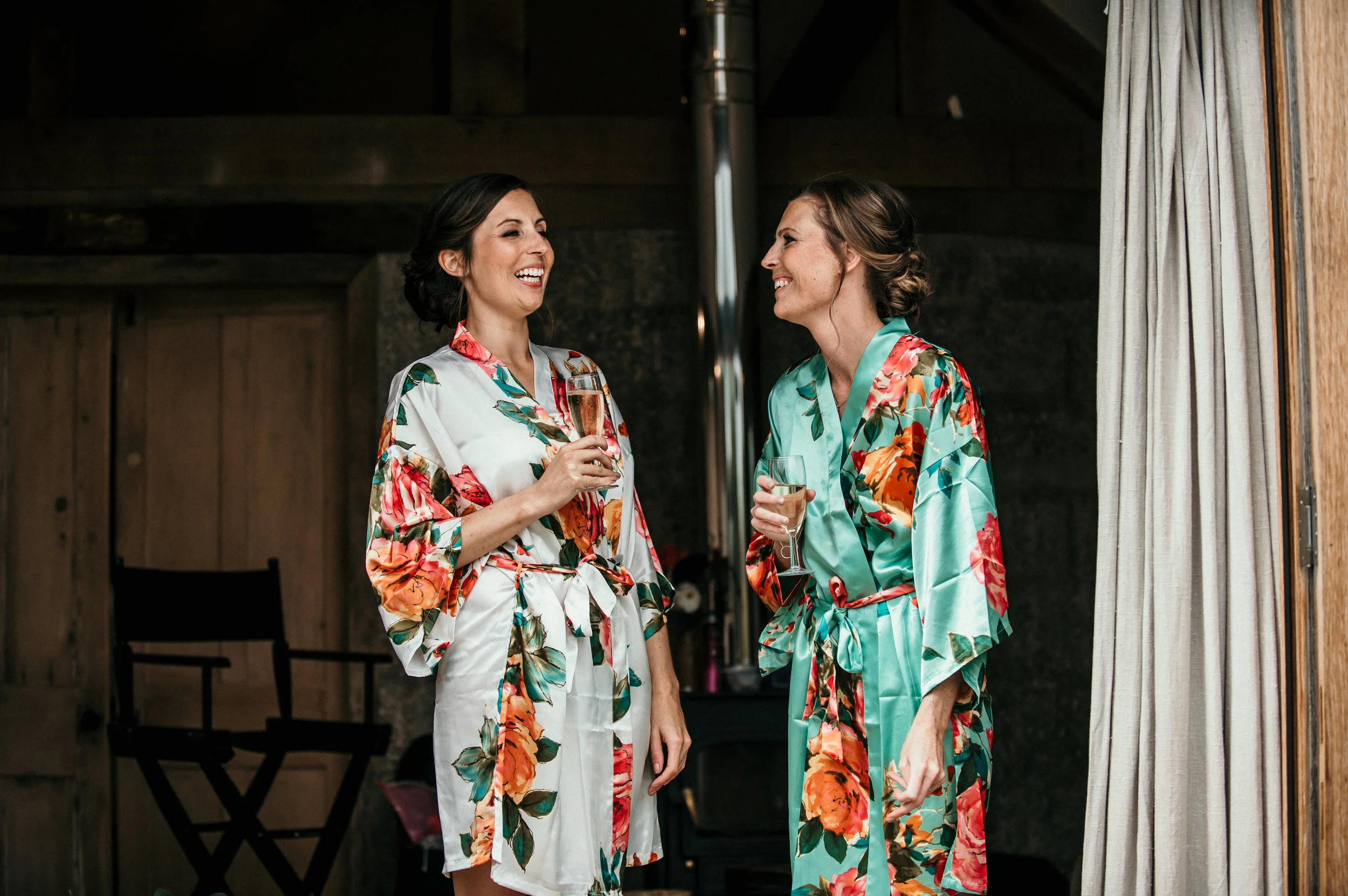 Two women in floral robes holding glasses of champagne, laughing and smiling indoors near a window with curtains.
