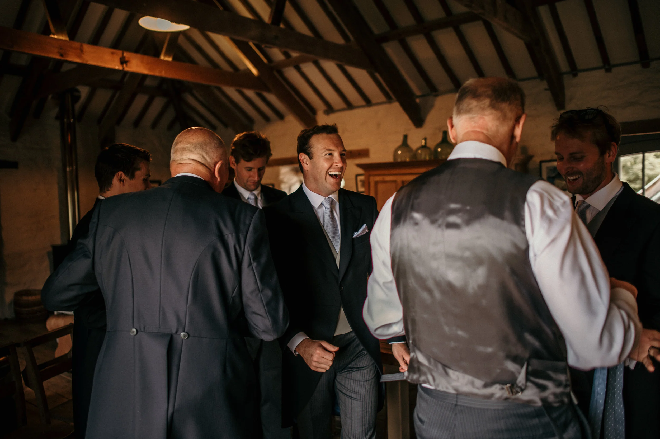 A group of men dressed in formal suits and waistcoats gather and chat joyfully in a rustic, warmly lit room with a high, wooden-beamed ceiling.