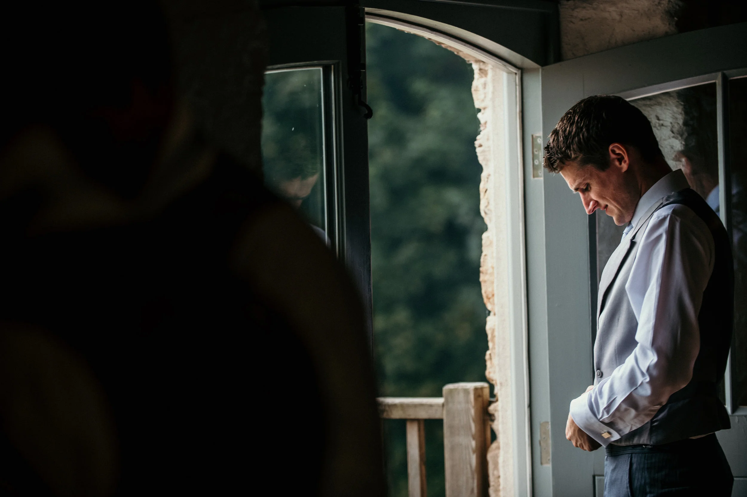 A man in formal attire, including a white dress shirt and vest, stands near an open door with light coming through. His head is bowed, and he appears to be adjusting his cufflinks or sleeves. The background outside the door is green and blurry.