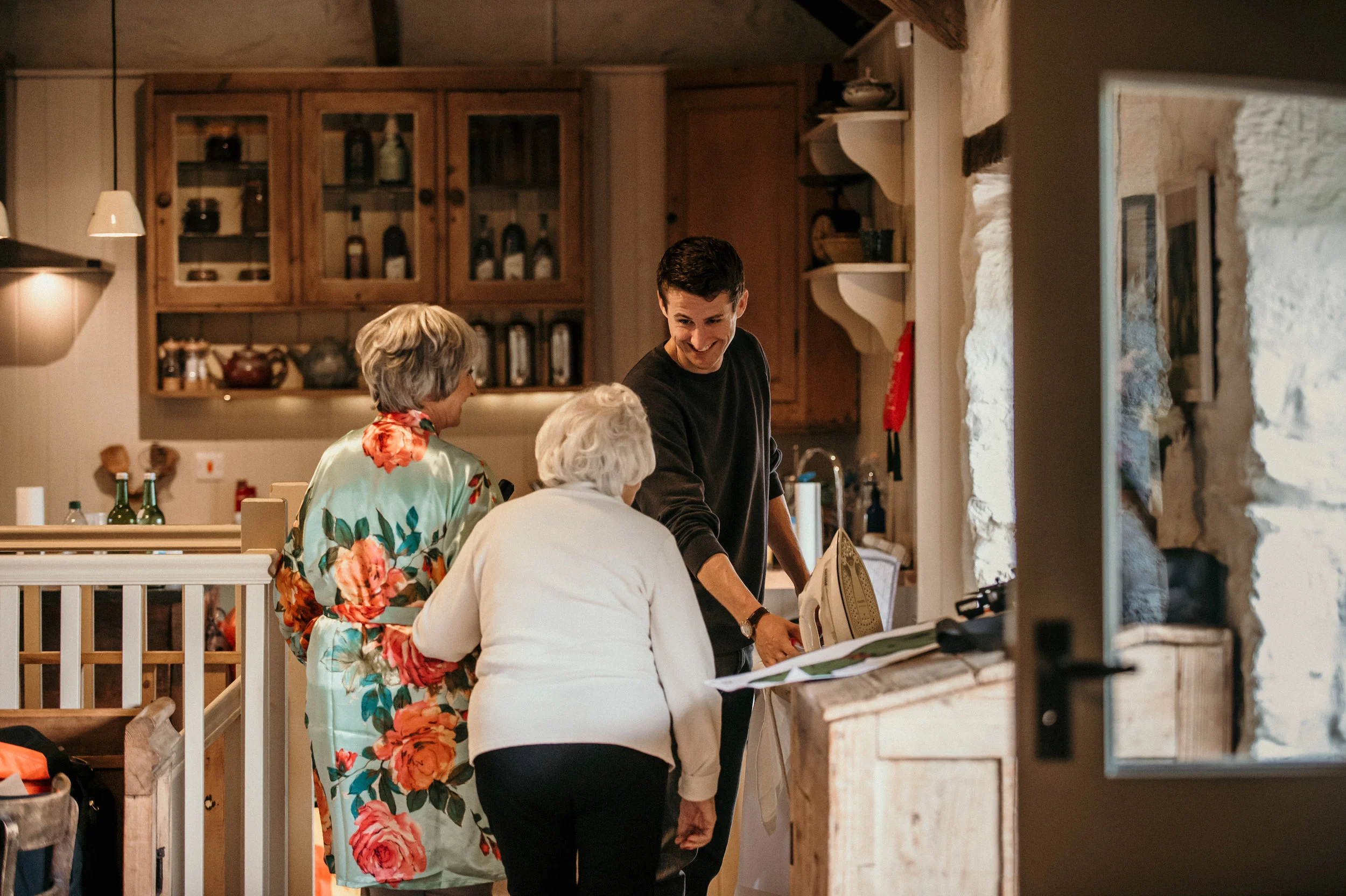 A young man doing laundry in a kitchen with two elderly women, one wearing a floral robe and the other a white sweater, sharing a moment together.