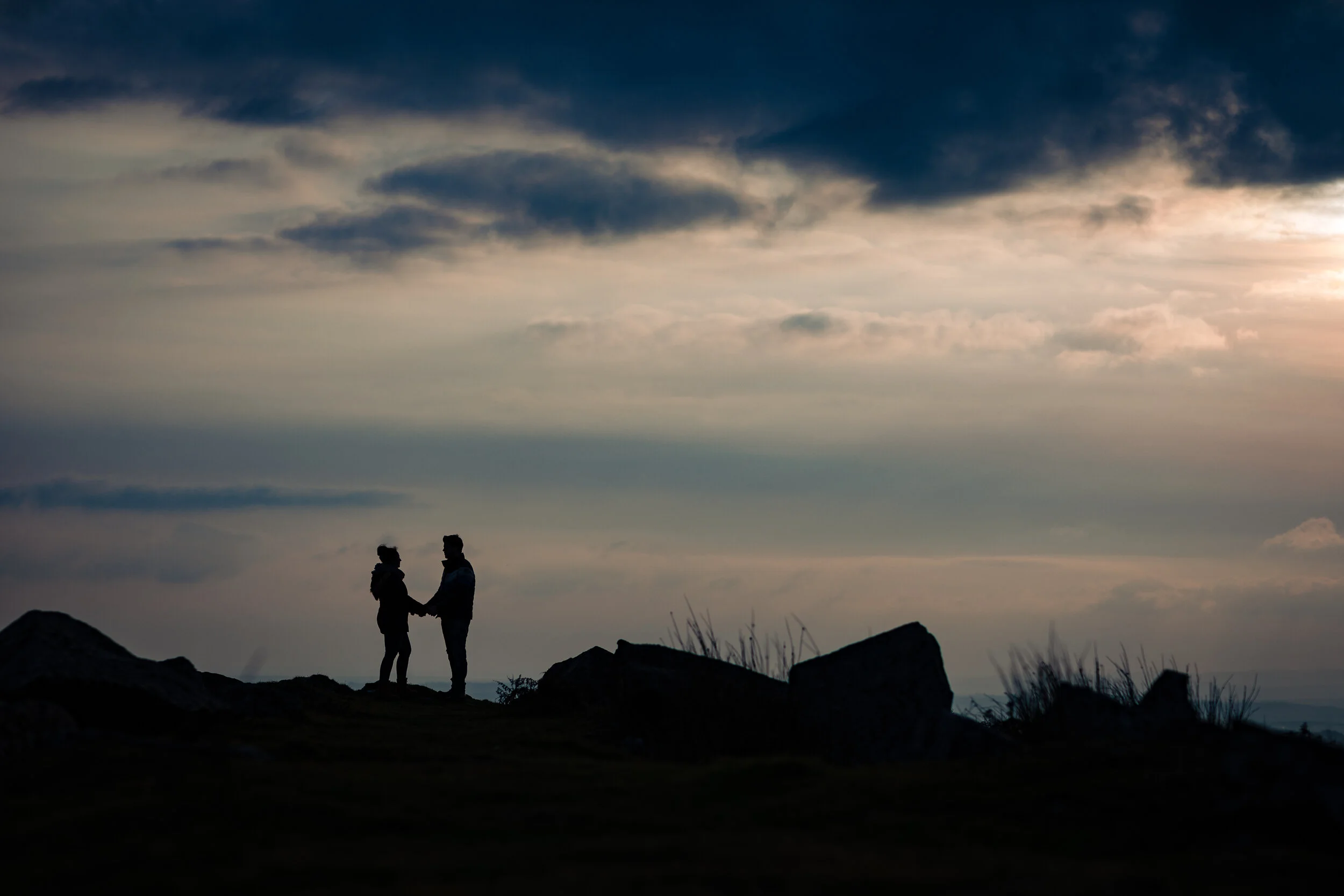Silhouette of a couple in Cornwall holding hands on a hilltop against a cloudy sky at sunset with ocean backdrop.