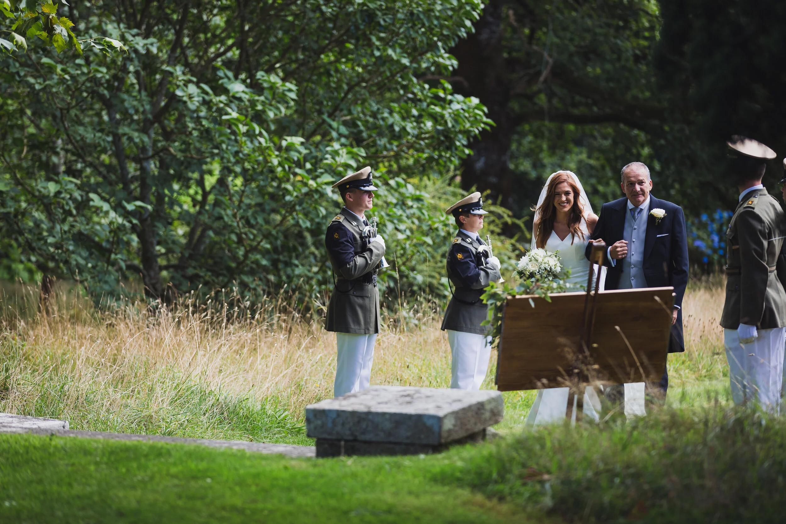 A couple getting married in an outdoor ceremony with military personnel, holding flowers and walking arm in arm, in a natural setting with trees and grass.