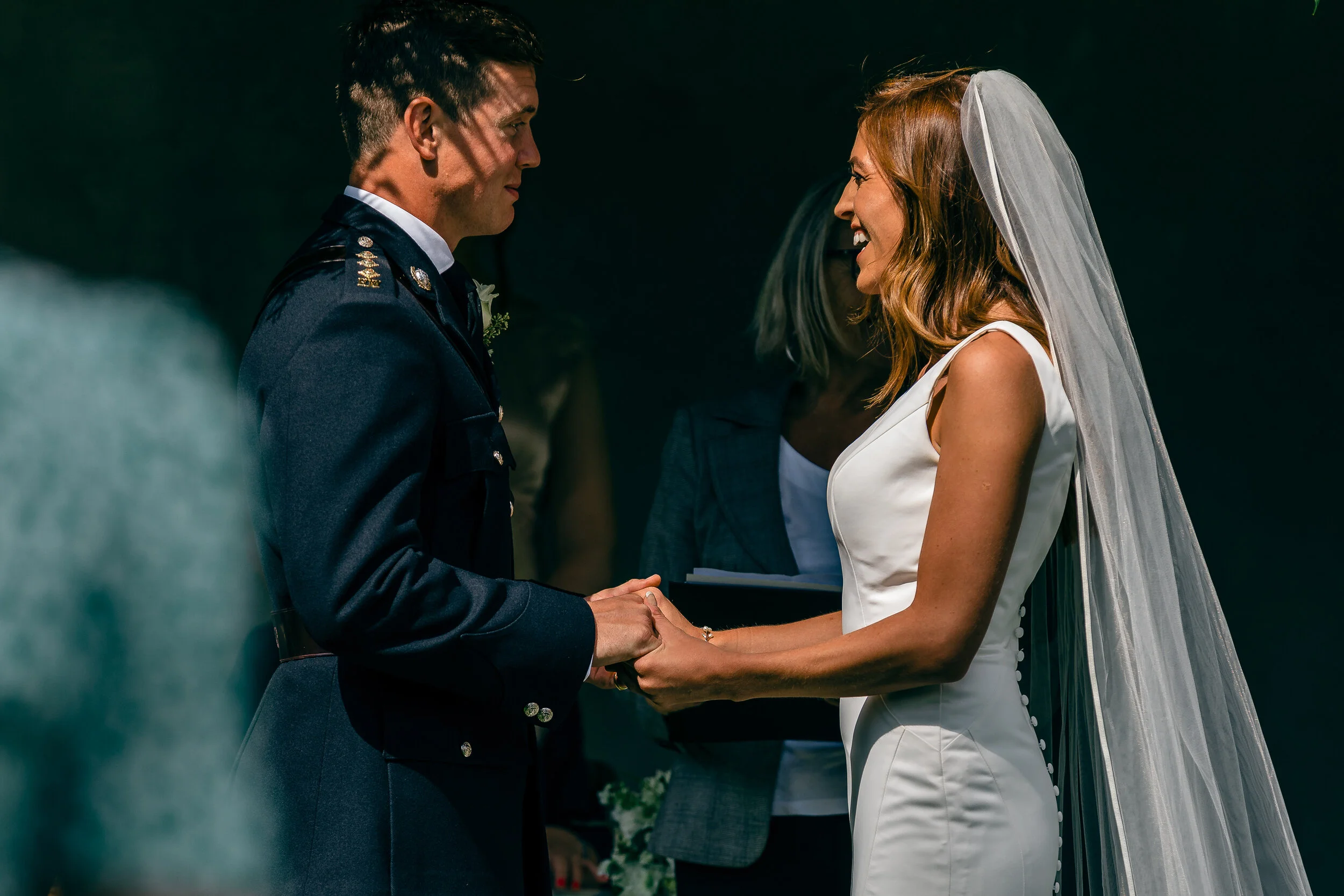A bride and groom holding hands and staring at each other during their wedding ceremony, with a officiant and a dark background.