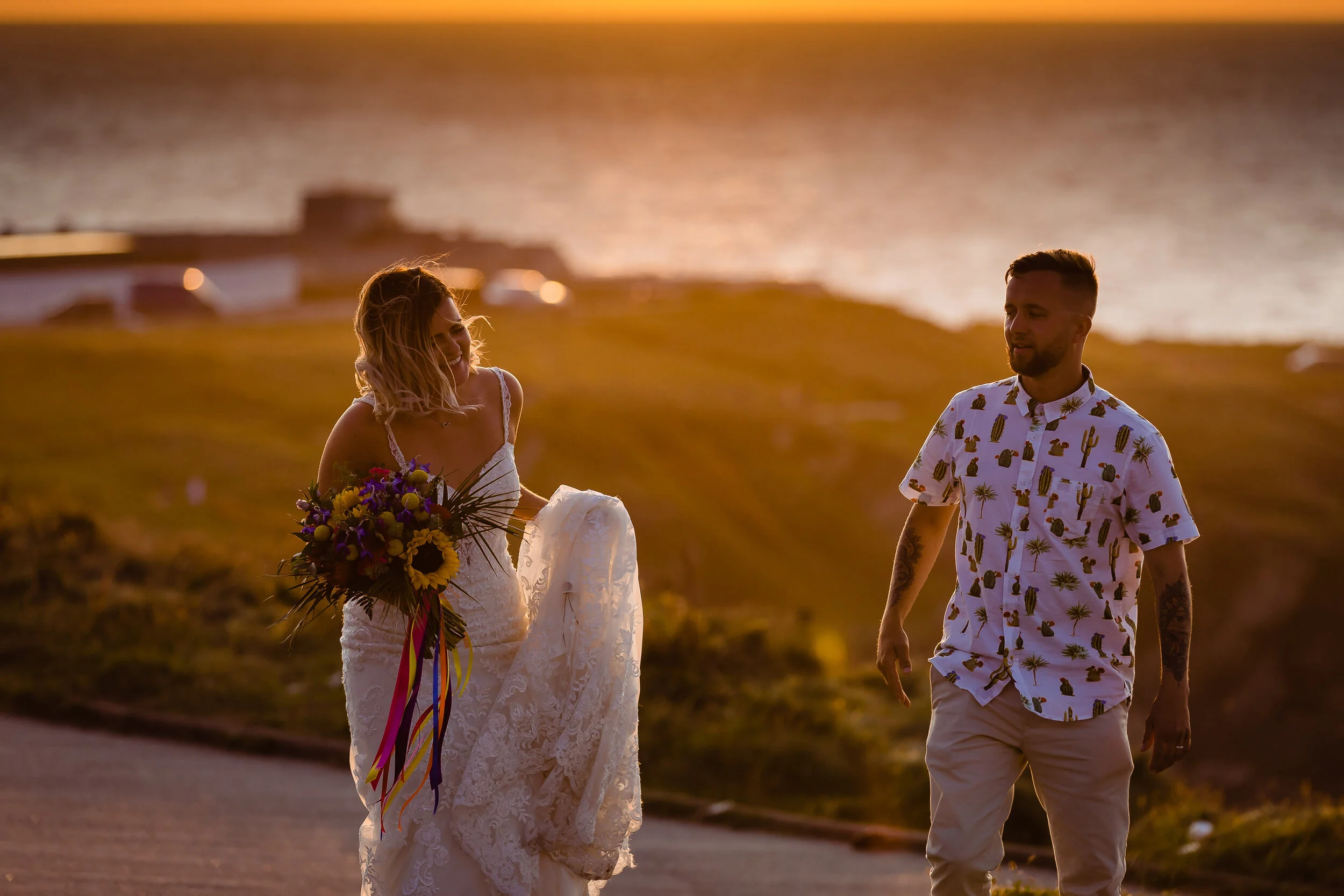 A woman in a white lace wedding dress holding a bouquet of colorful flowers walks along a scenic coastal path at sunset, with a man in a short-sleeved shirt with a cactus and palm tree pattern walking beside her.