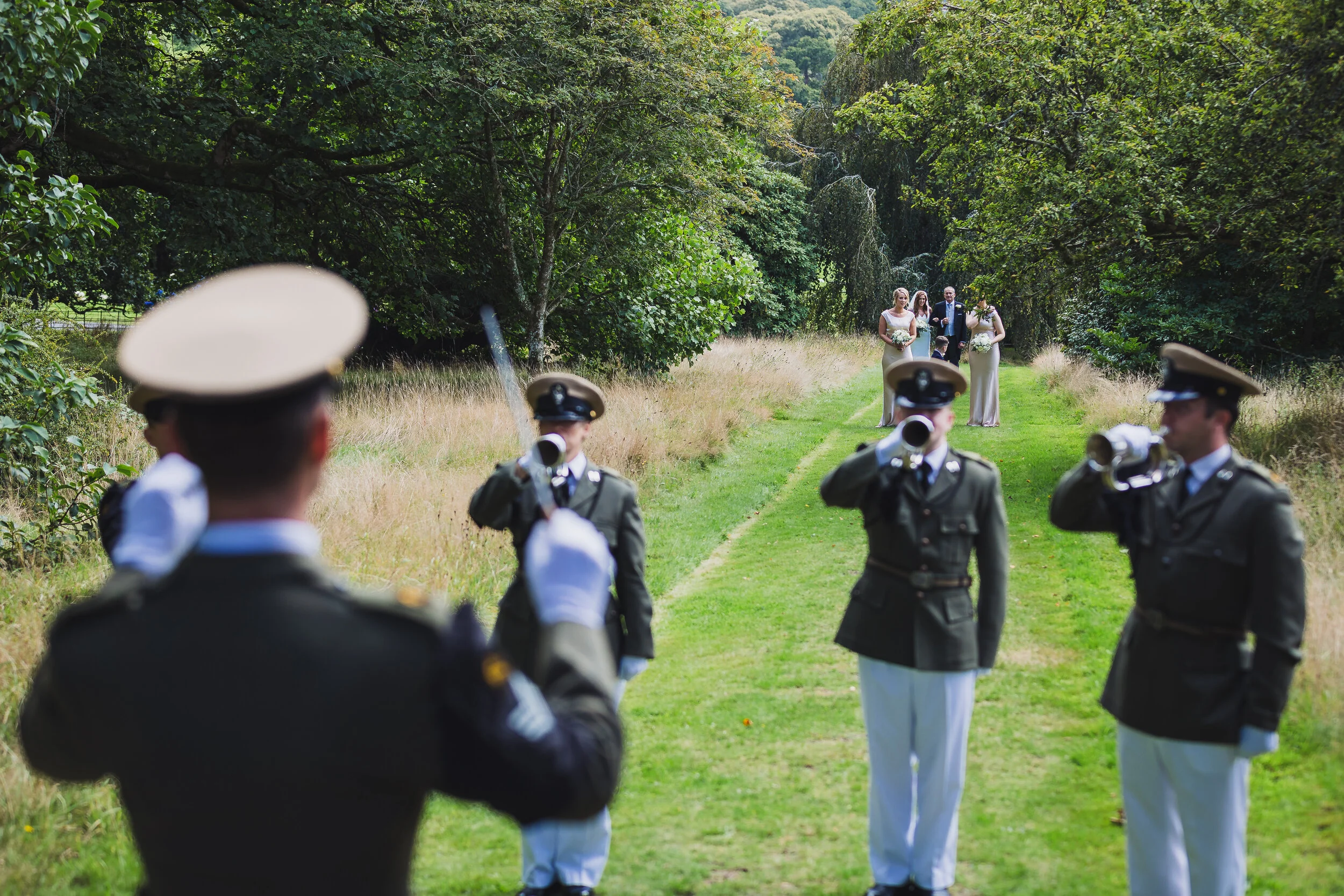 Military personnel in uniform standing in formation on a grassy path, saluting, with a wedding party walking towards them in the background in a lush green outdoor setting.