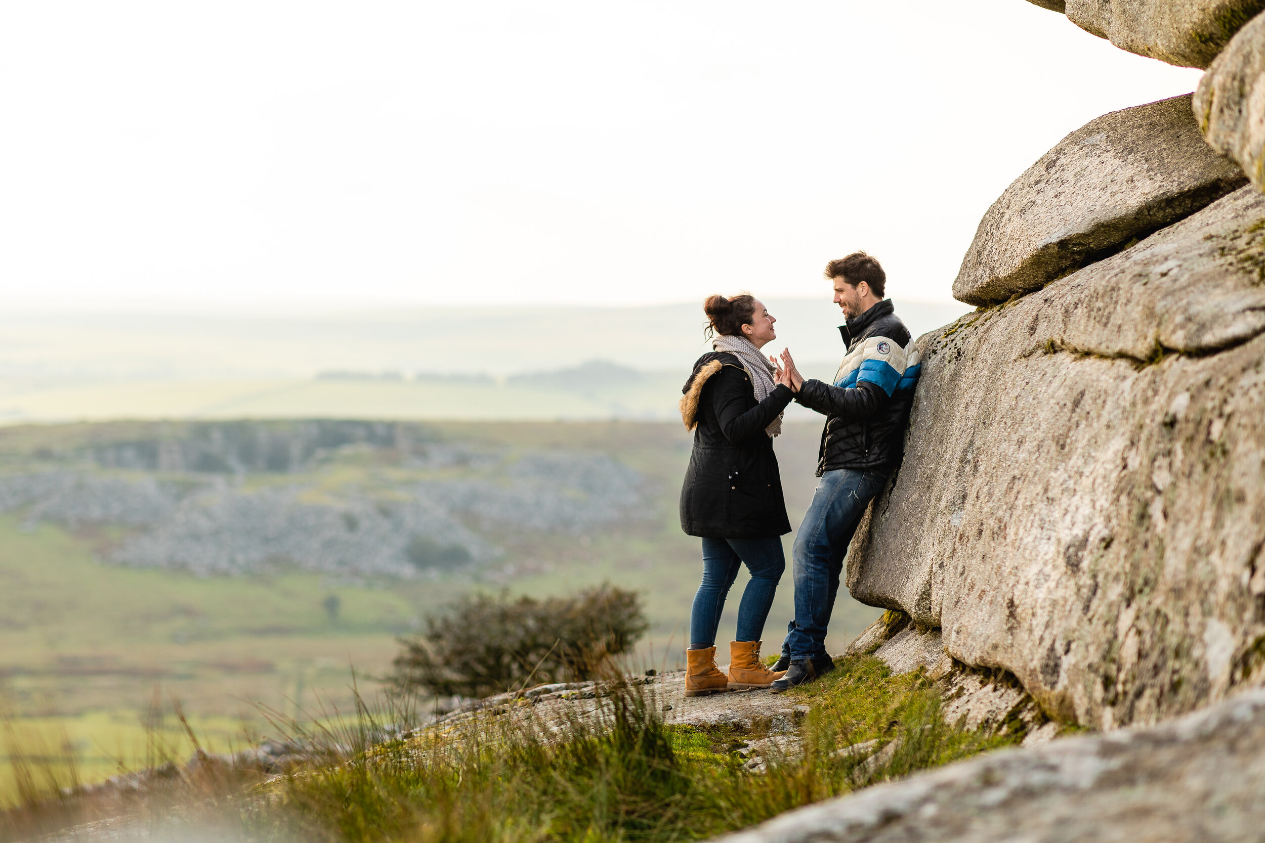 A couple smiling and touching hands near a large stone wall outdoors in a countryside setting with rolling hills and green fields in the background.