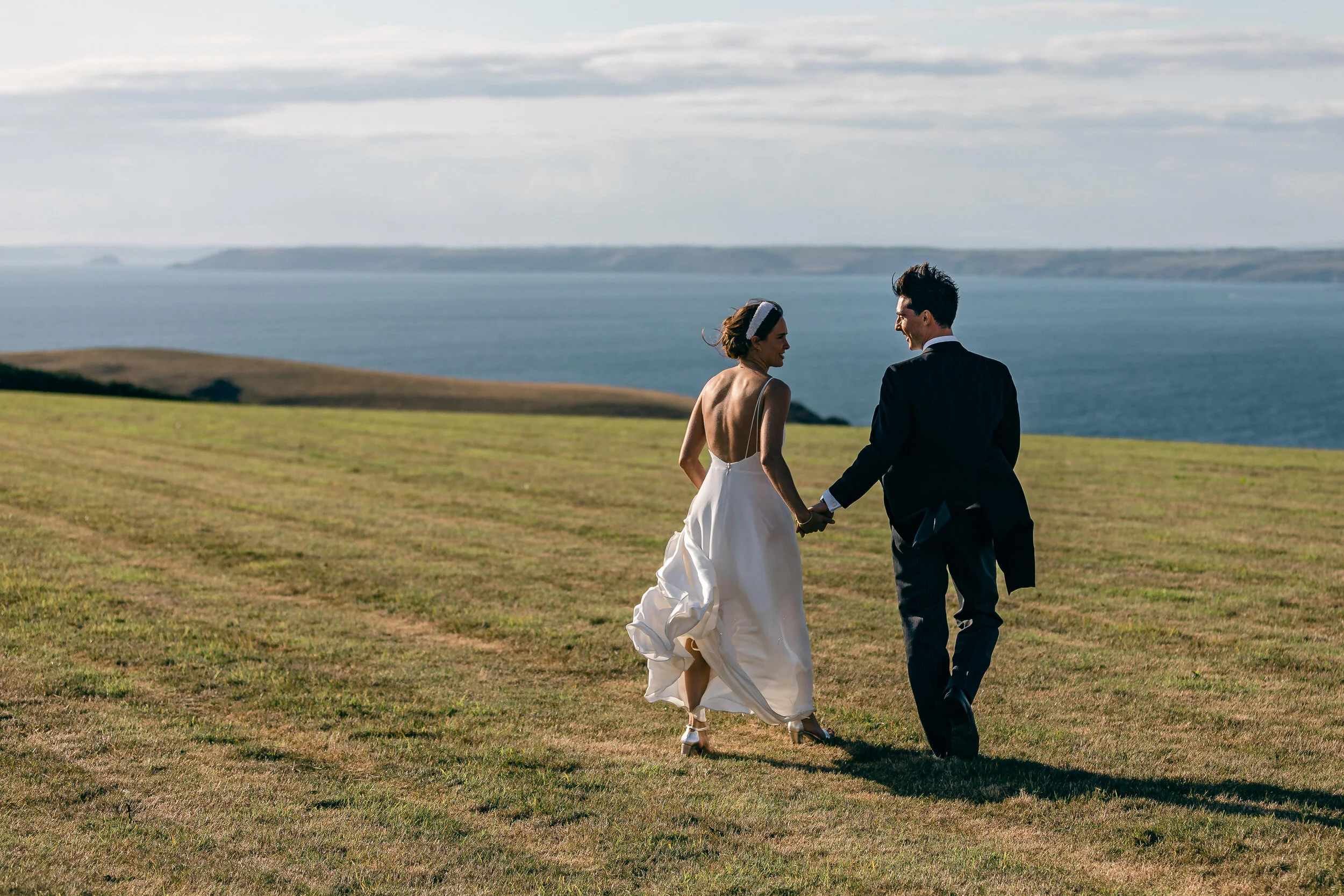 A bride and groom walking hand in hand across a grassy field with the ocean and cloudy sky in the background.