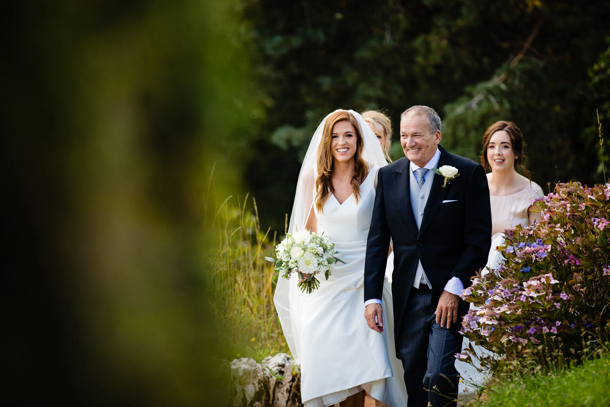 A bride in a white wedding dress and a man in a dark suit walking outdoors with two women behind them, one holding a bouquet, surrounded by greenery and flowers.