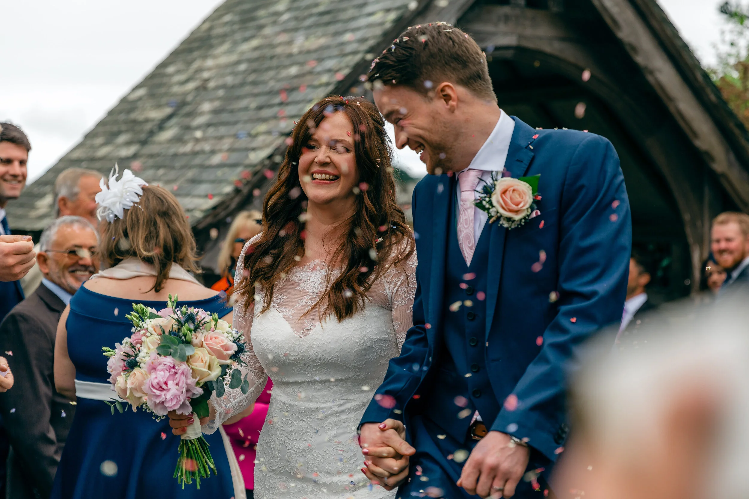 A bride and groom holding hands and smiling during their wedding celebration, surrounded by guests and colorful confetti outside a rustic building.